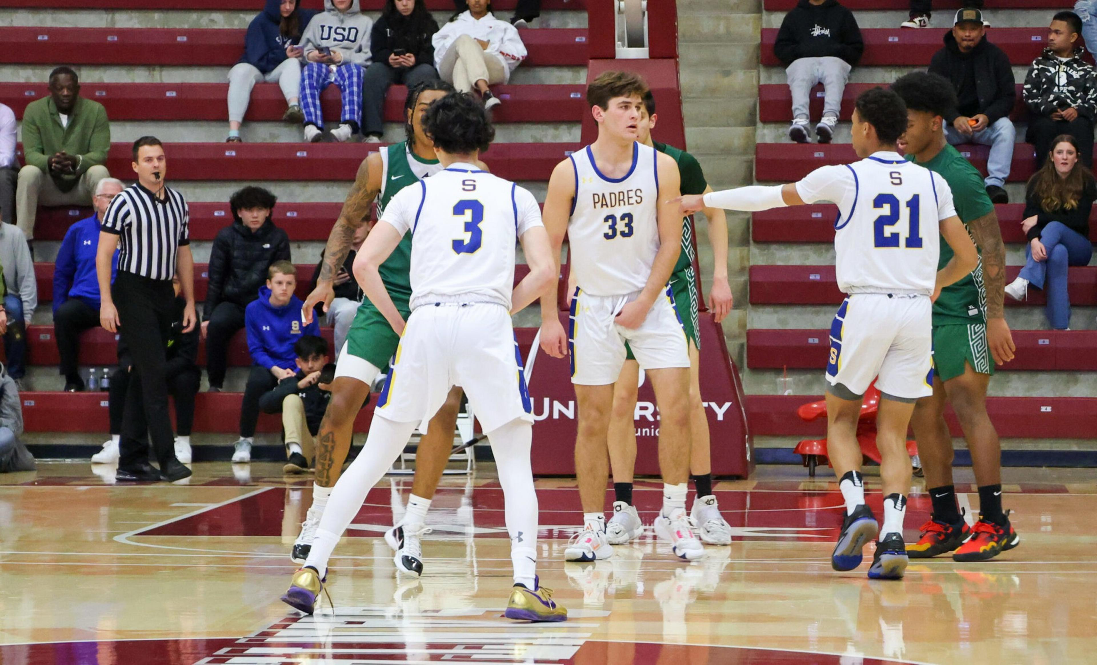 Three basketball players in white jerseys with "Padres" face off against players in green jerseys on a gym court, with spectators and a referee nearby.