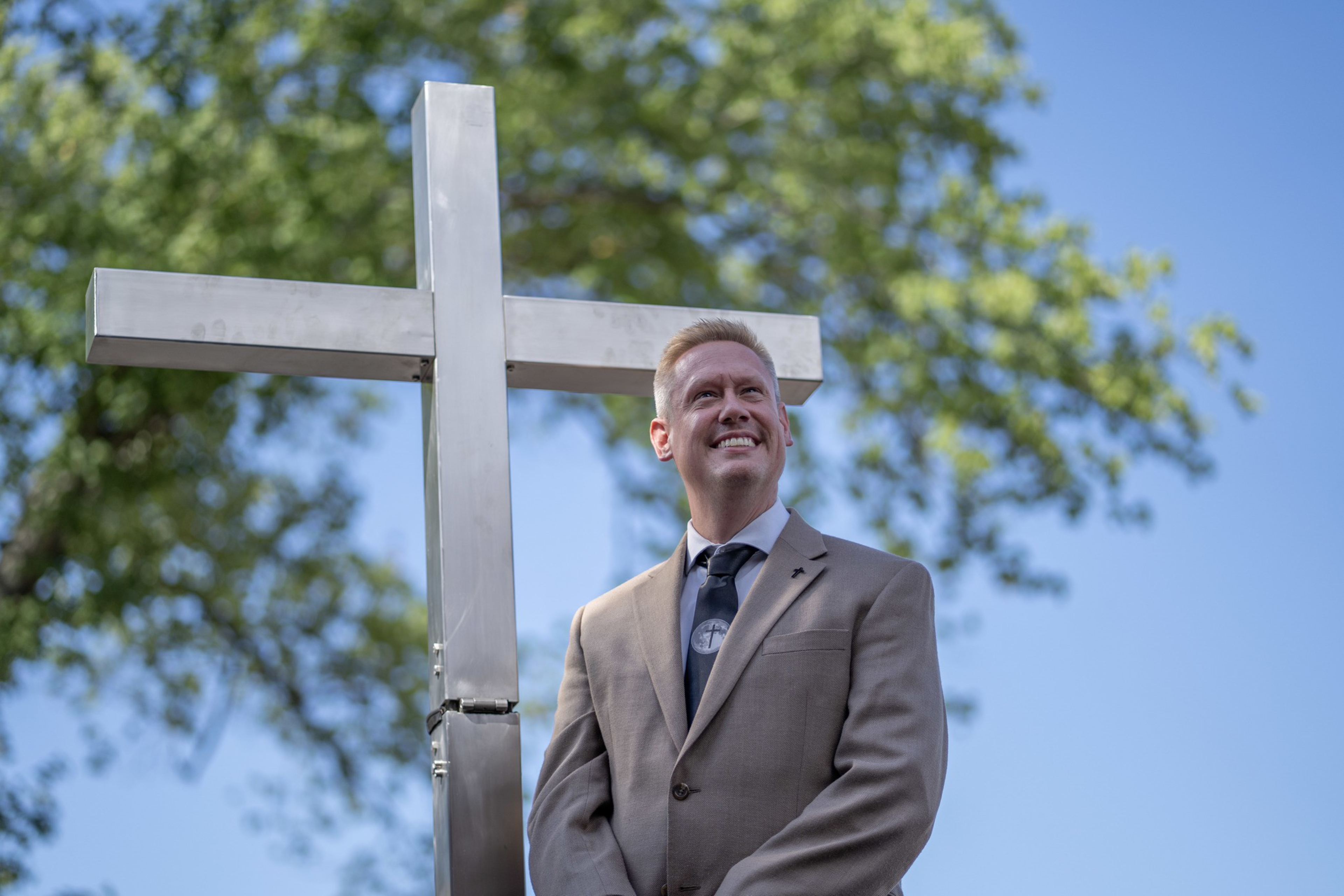 A man in a tan suit and dark tie stands smiling beside a large metallic cross with trees and blue sky in the background.