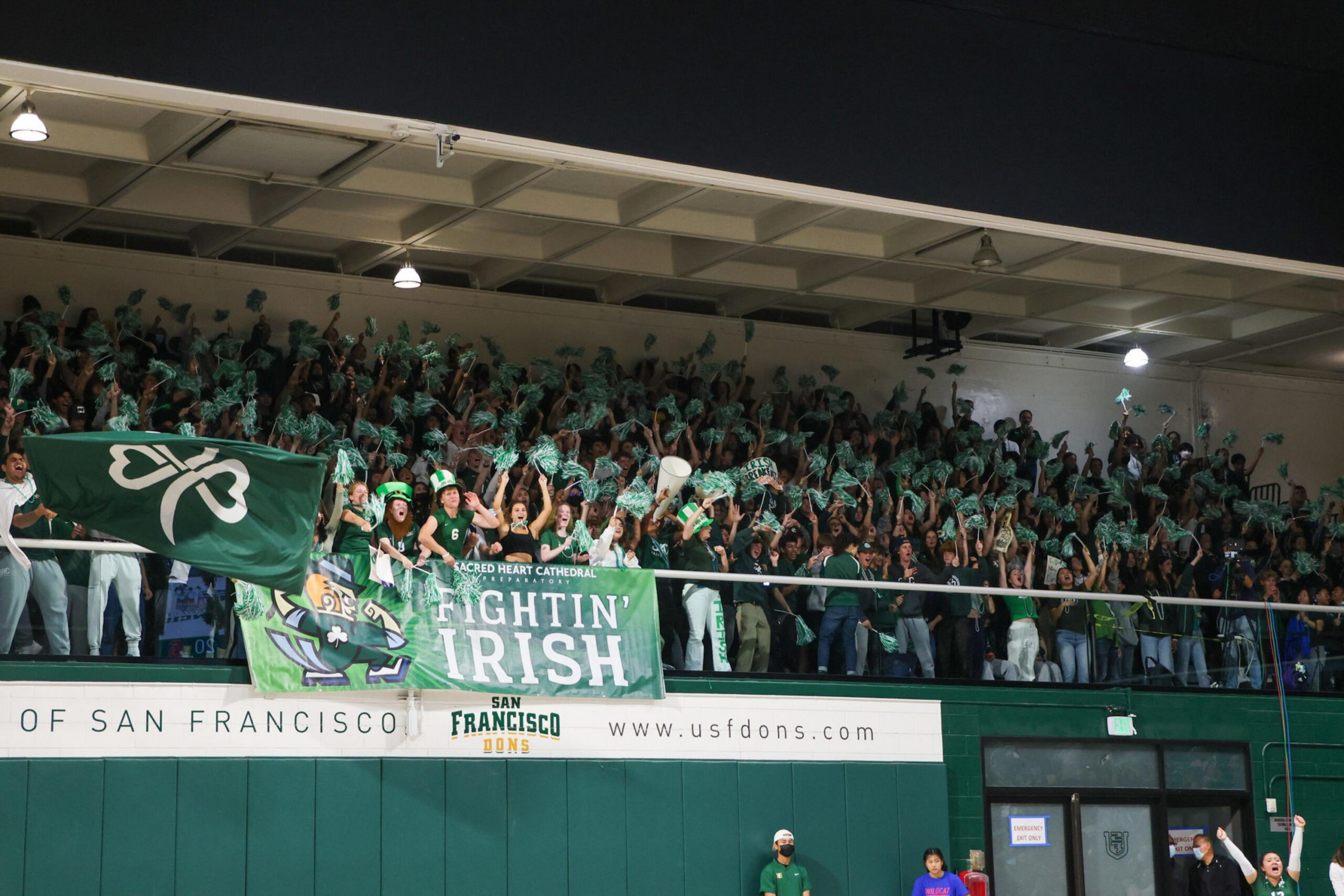 A large crowd of fans wearing green cheer enthusiastically behind banners that say "FIGHTIN' IRISH" and "SAN FRANCISCO DONS" in a gym.