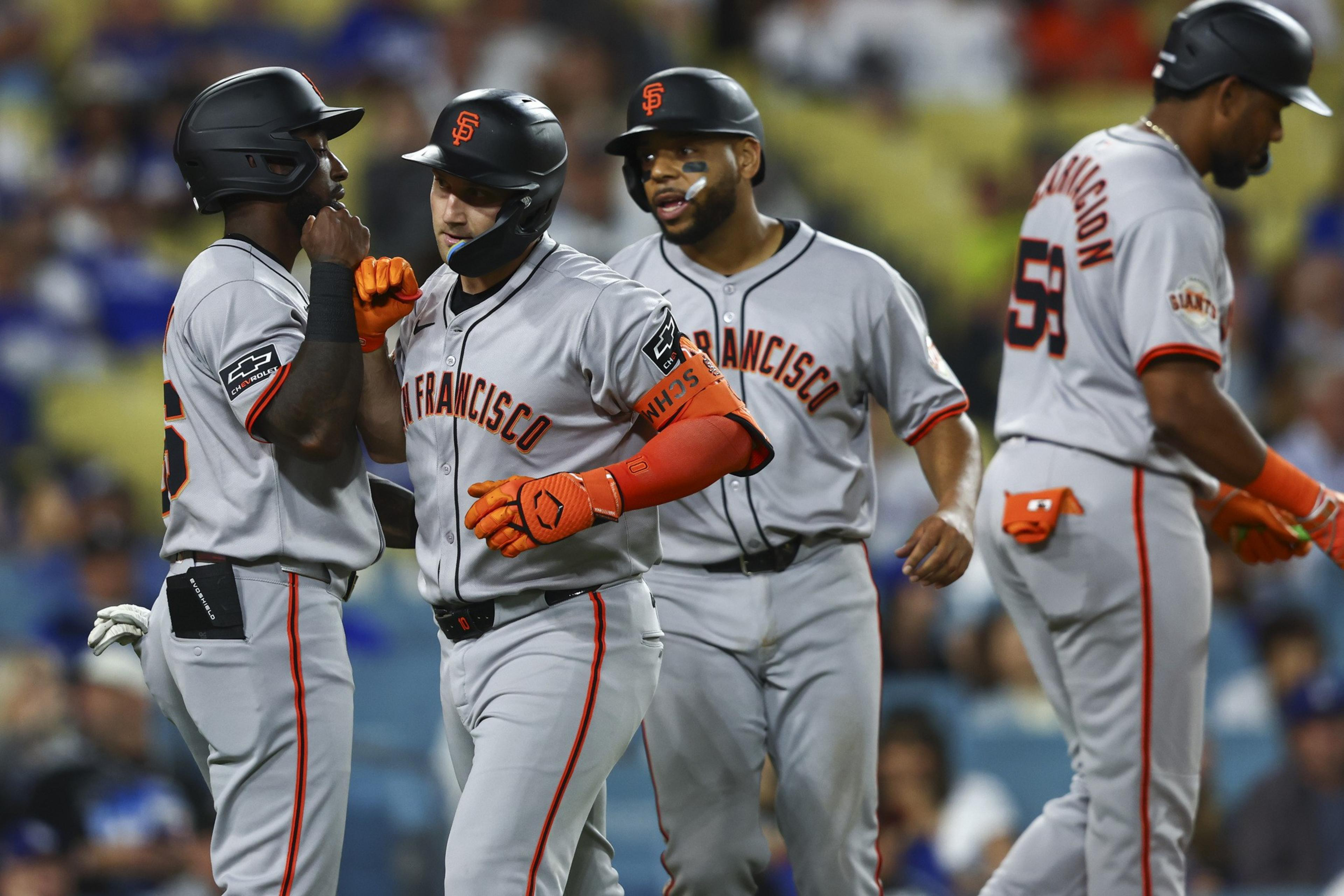 Four baseball players in gray &quot;San Francisco&quot; uniforms celebrate on the field, wearing black helmets and orange gloves, with a blurred crowd in the background.