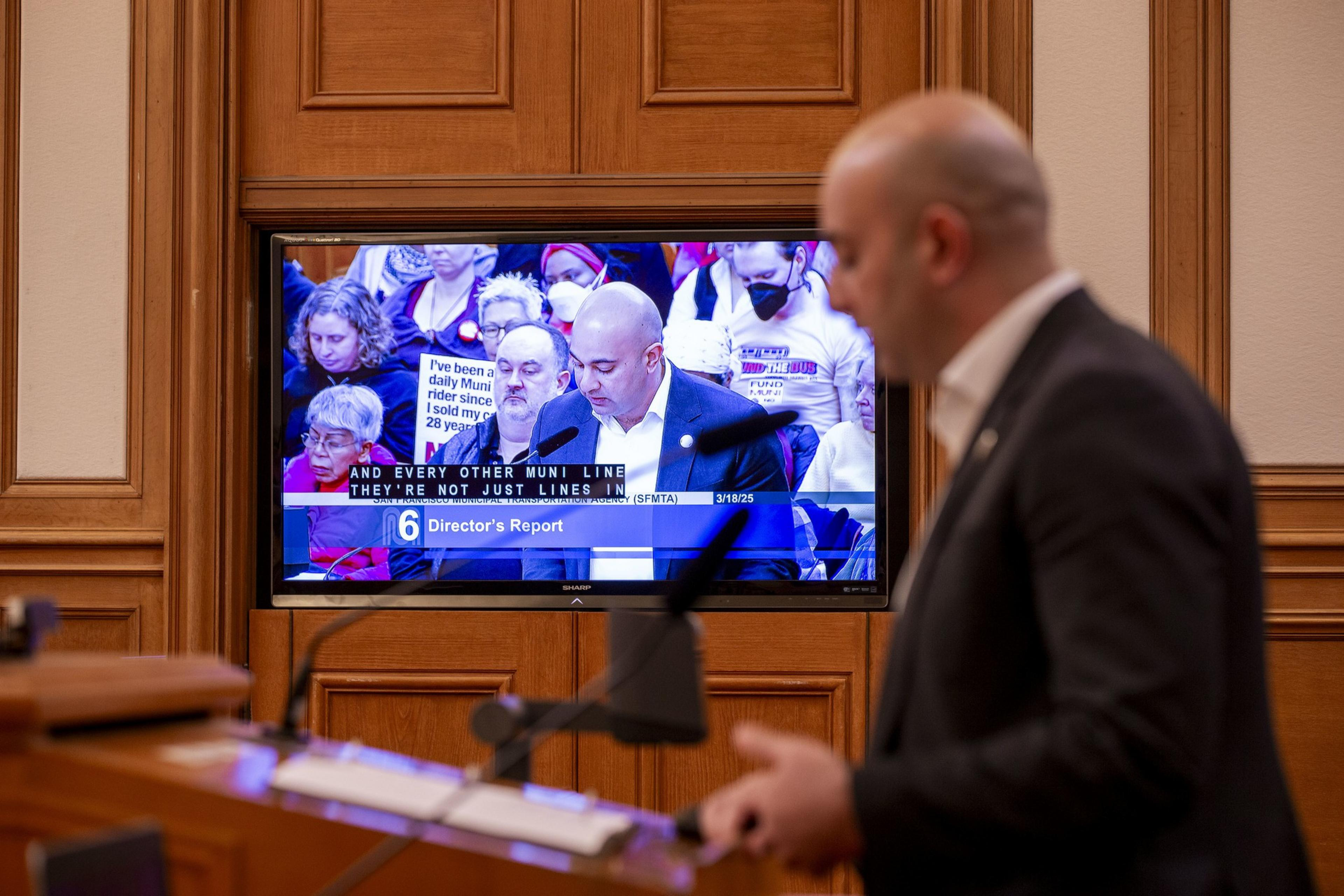 A man stands at a podium in focus, while a screen shows another man speaking at a meeting, with attendees seated. Captions display on the screen.