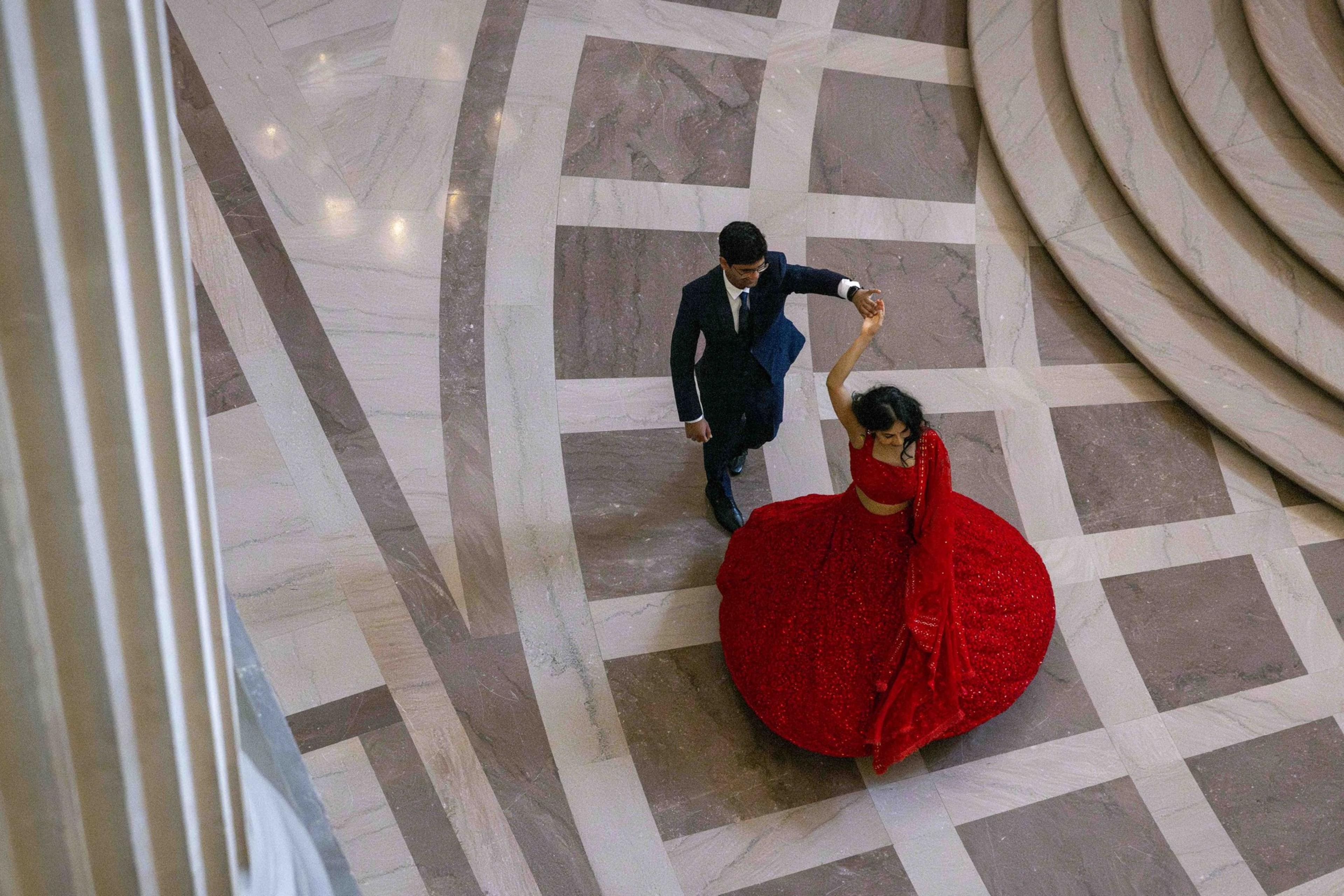 A couple dances on a patterned marble floor. The man in a dark suit twirls the woman, who's wearing a vibrant red dress, creating a dynamic swirl.