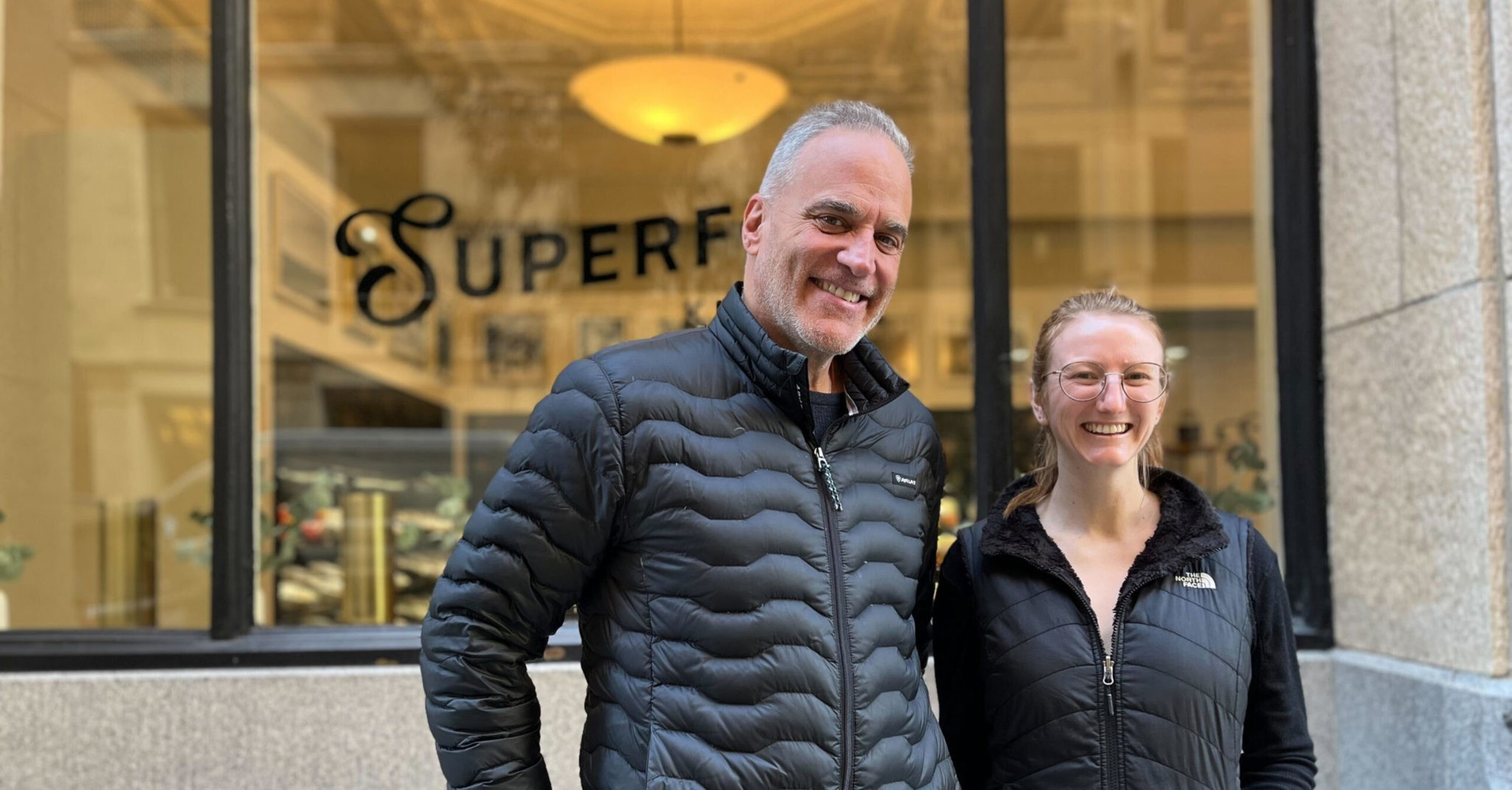 A man and a woman, both wearing black jackets, stand smiling outside a building. The word "SUPERF" is partially visible on the window behind them.