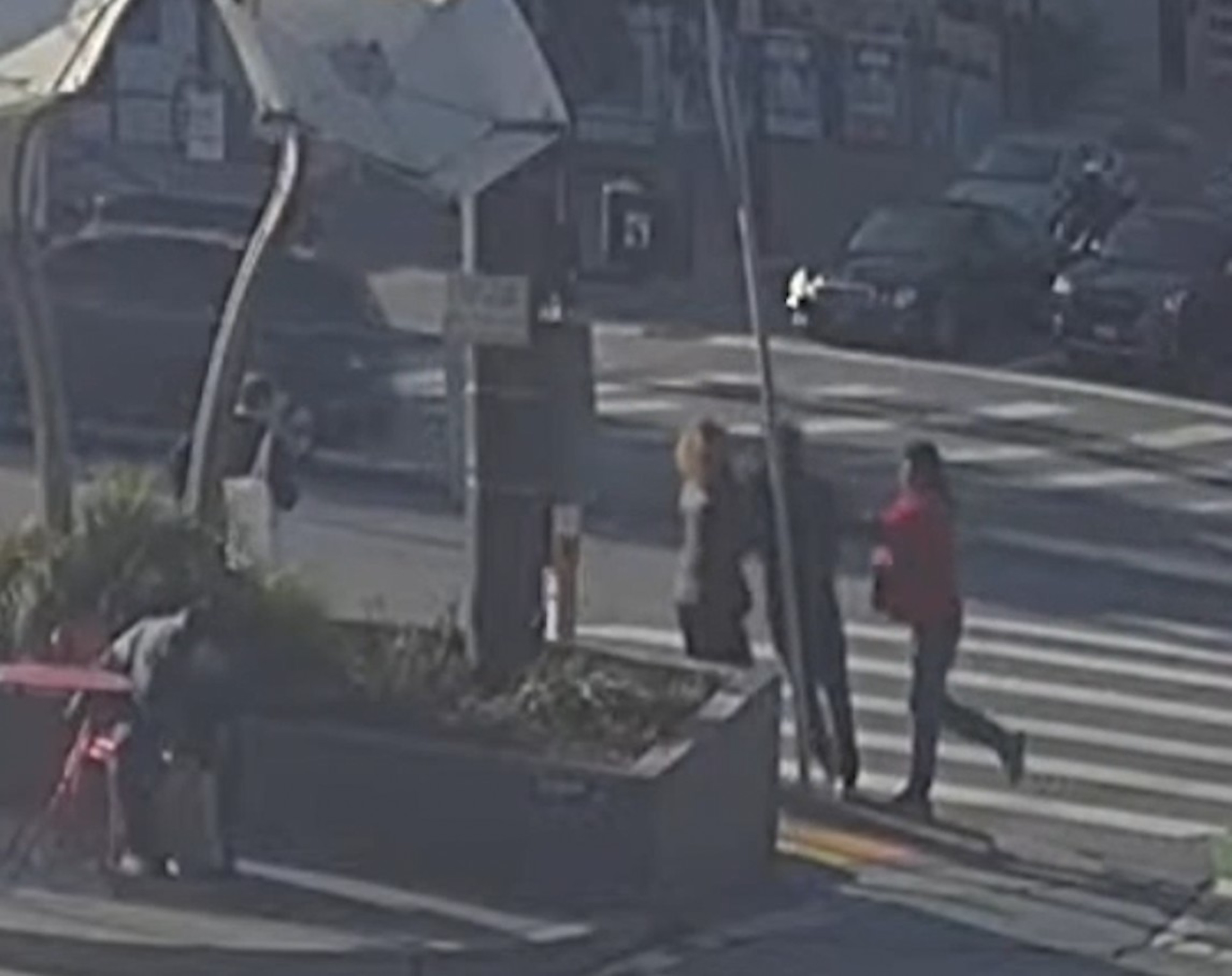 Three people are crossing a street at a crosswalk. A fourth person is hunched over near a red table. Cars and other pedestrians are visible in the background.
