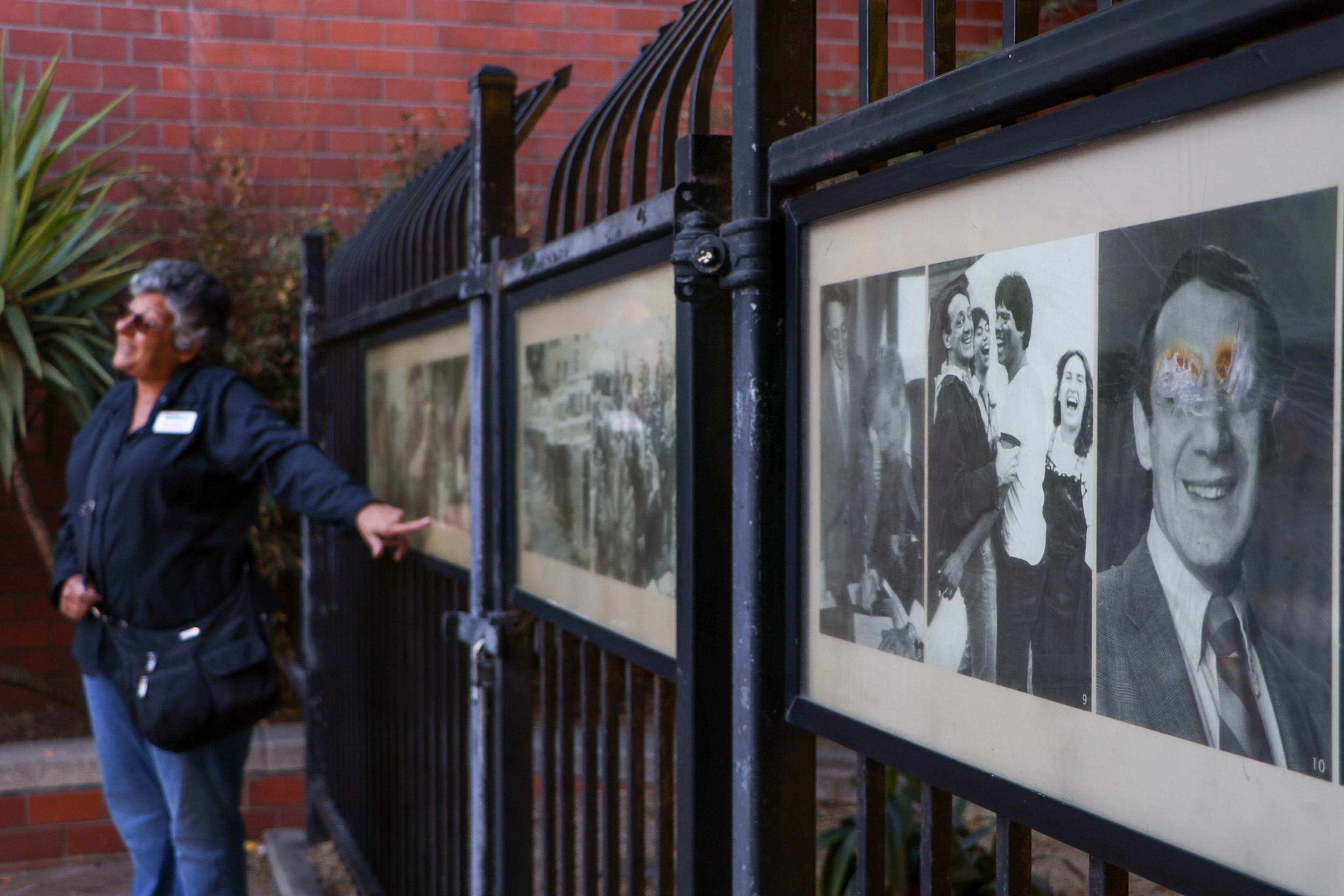 A woman stands near a black metal fence with framed black-and-white photos, pointing at one while wearing sunglasses and casual clothes.