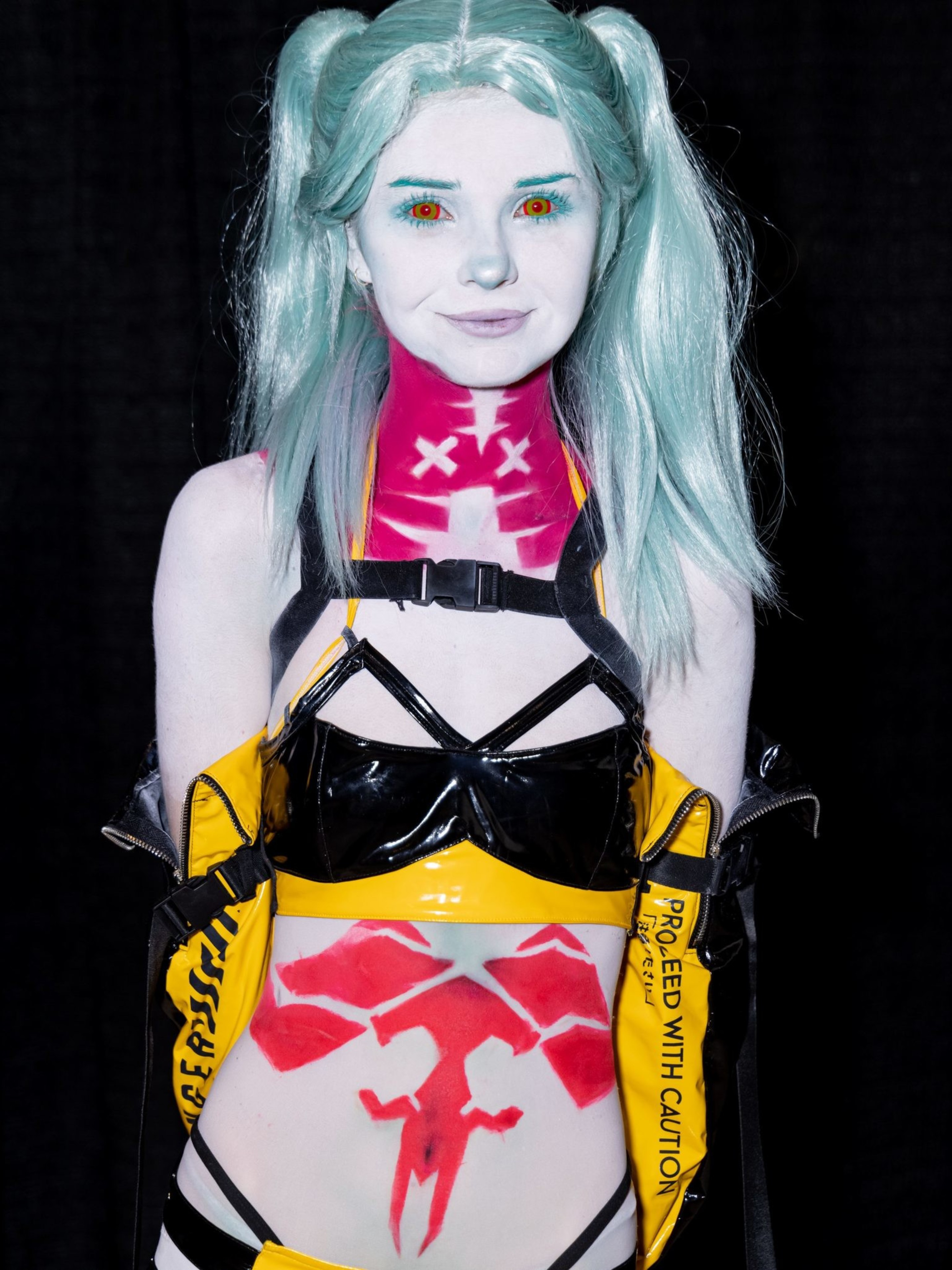 A person with light turquoise hair and red eyes wears elaborate black and yellow clothing with futuristic designs and symbols, including red markings.