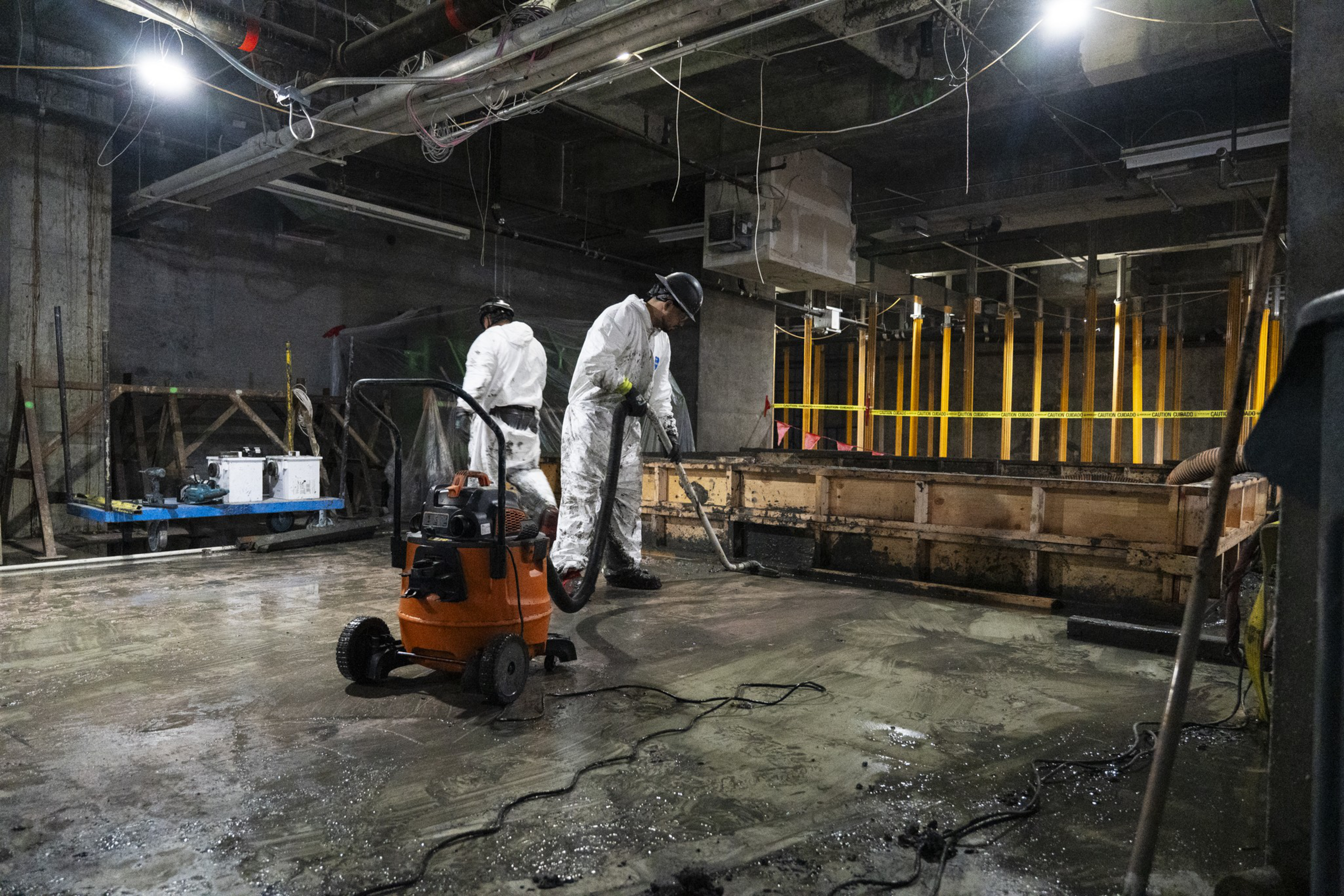 Two workers in white protective suits and helmets clean a wet concrete floor in a dimly lit construction site using a vacuum machine.
