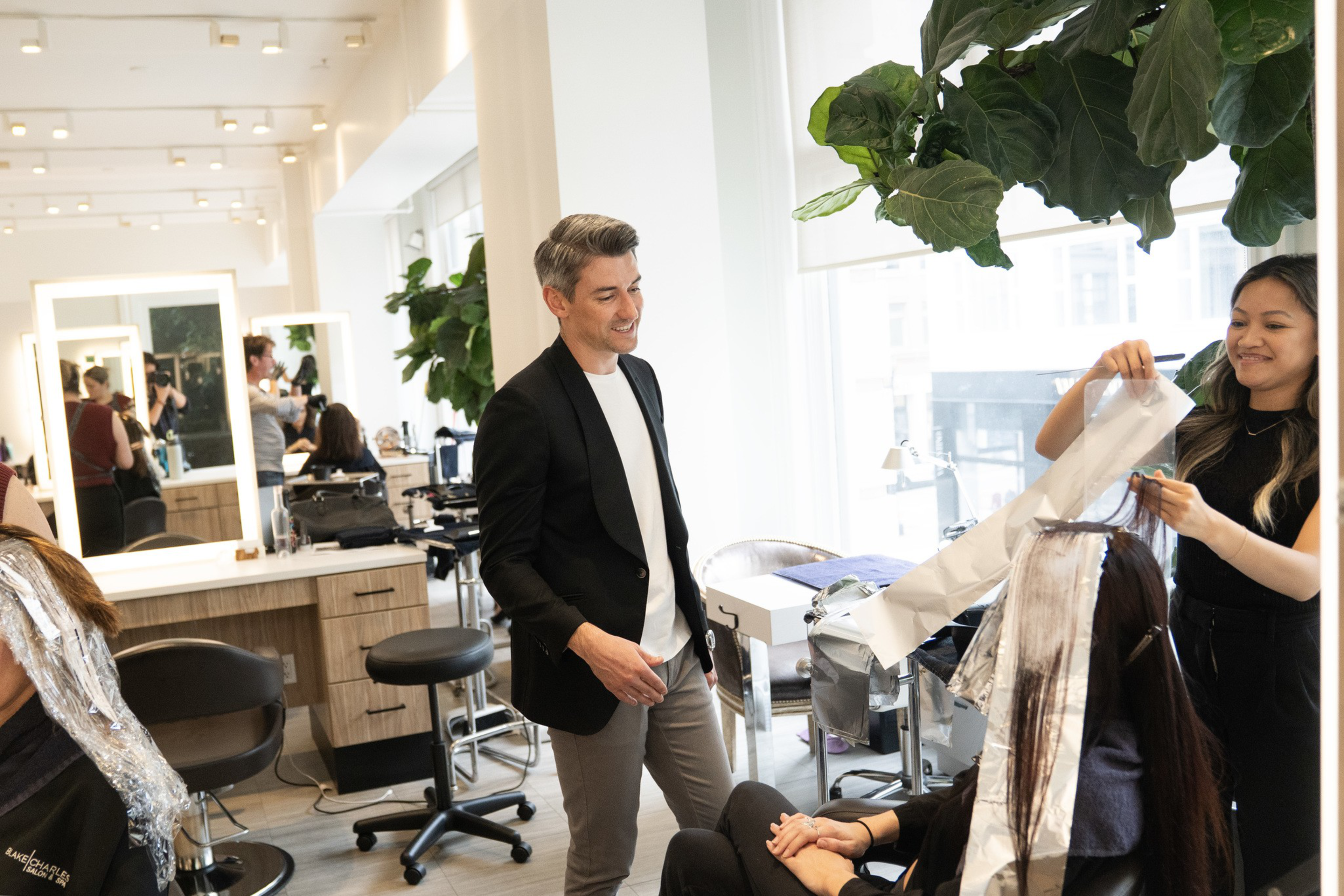 A hairstylist applies foils to a seated woman's hair while a man in a black blazer and white shirt smiles nearby in a bright salon.