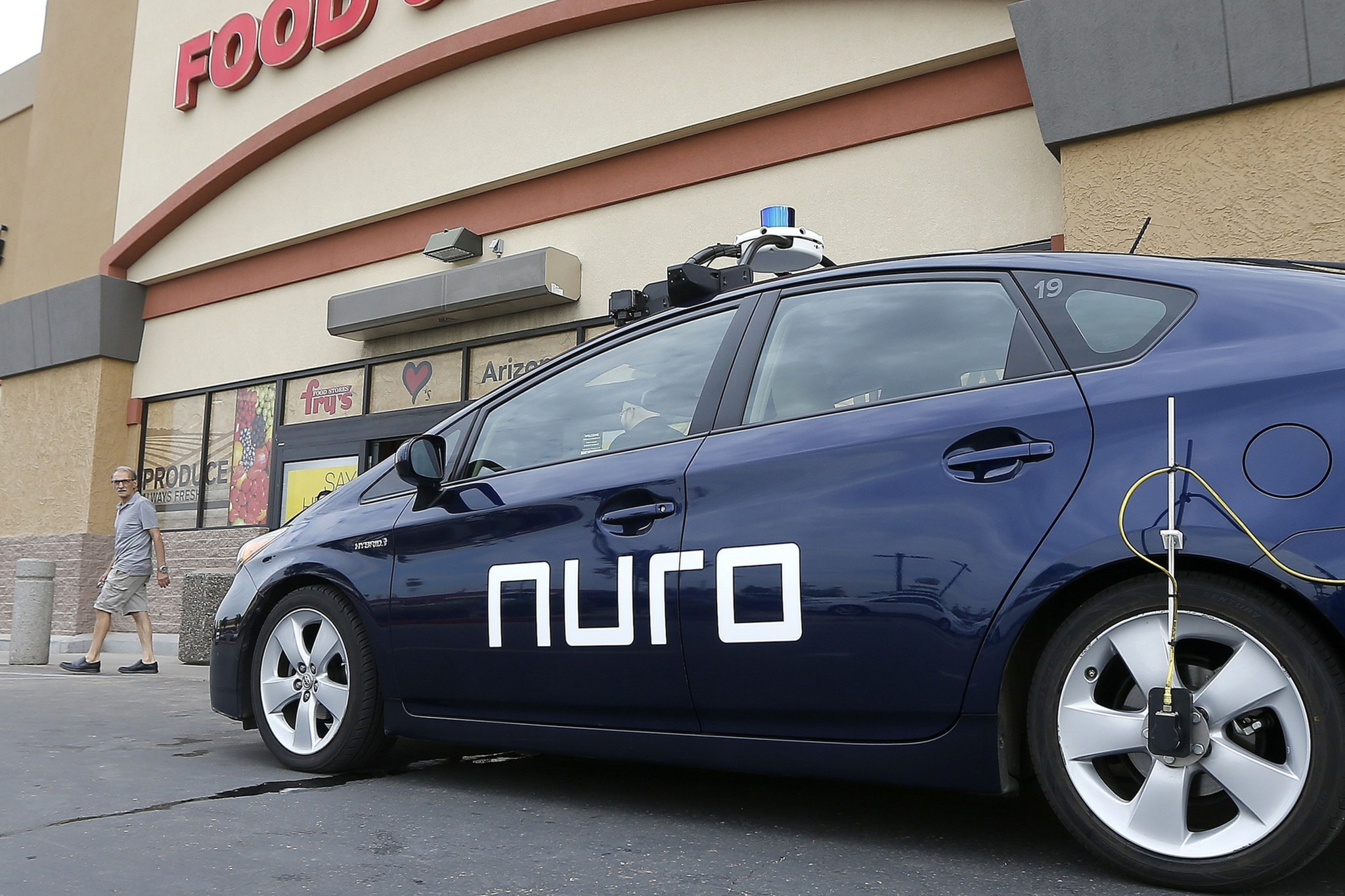 A dark blue self-driving car with "nuro" written on the side is parked outside a store with a partial sign reading "FOOD &". A man walks nearby.