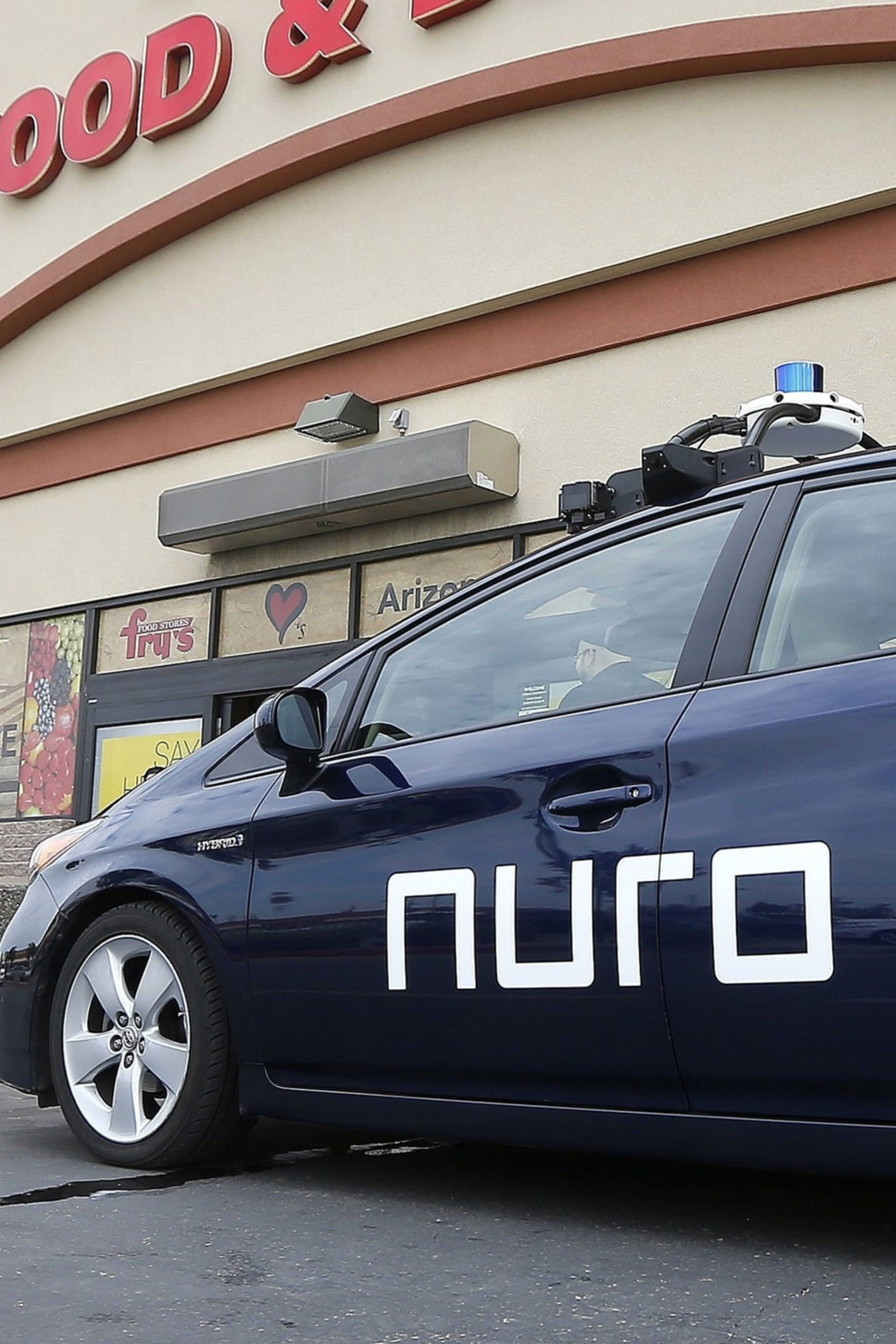 A dark blue self-driving car with "nuro" written on the side is parked outside a store with a partial sign reading "FOOD &". A man walks nearby.