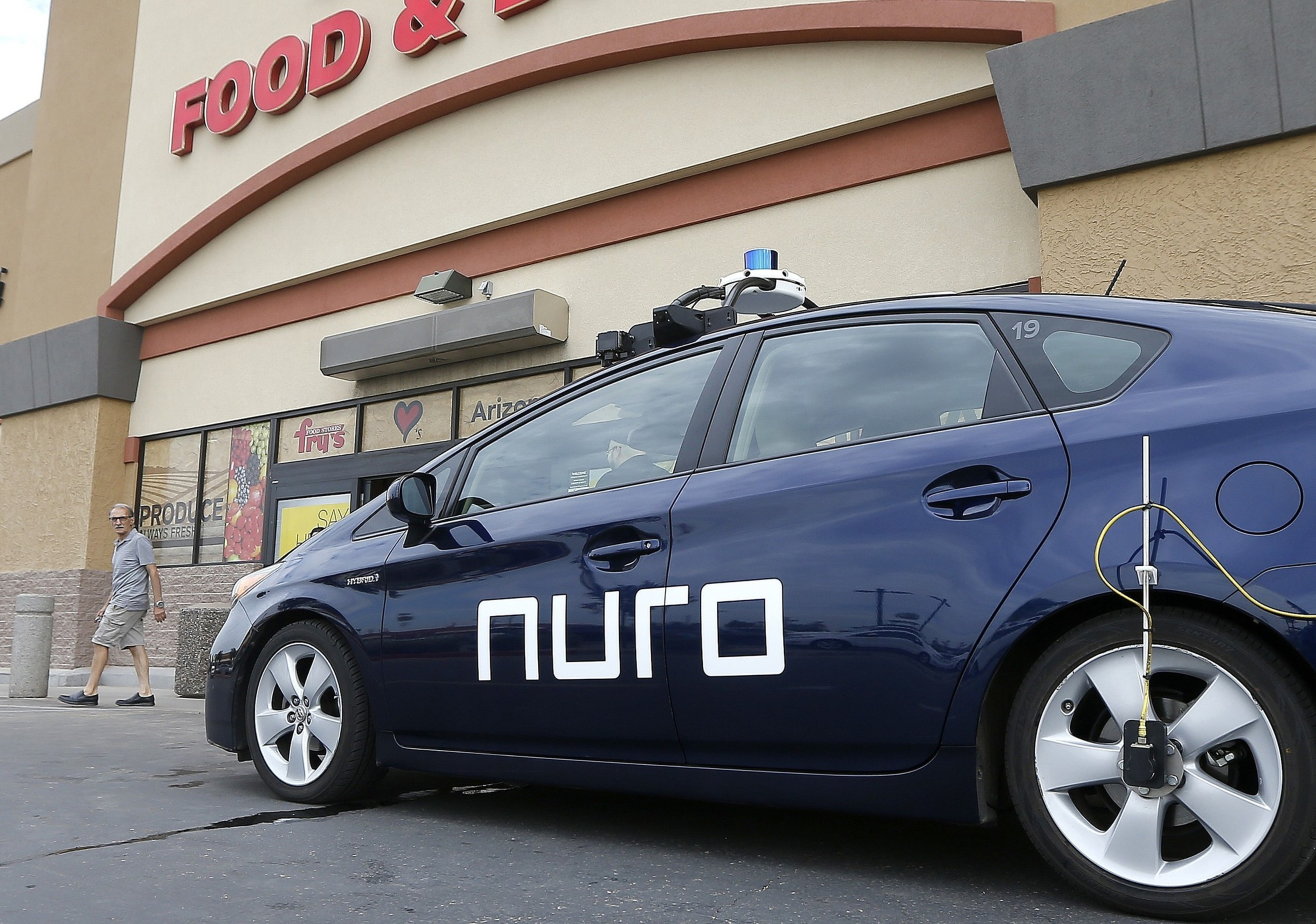 A dark blue self-driving car with "nuro" written on the side is parked outside a store with a partial sign reading "FOOD &". A man walks nearby.