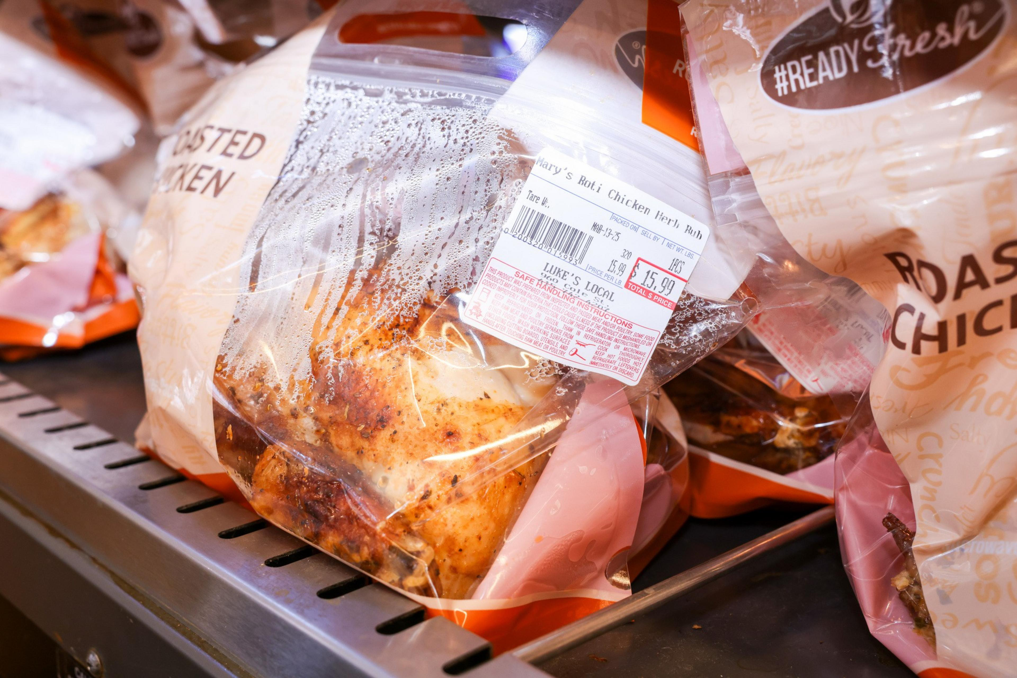 The image shows packaged roasted chickens in clear bags with labels. They are placed on a display rack, priced at $15.99, with condensation inside the bags.
