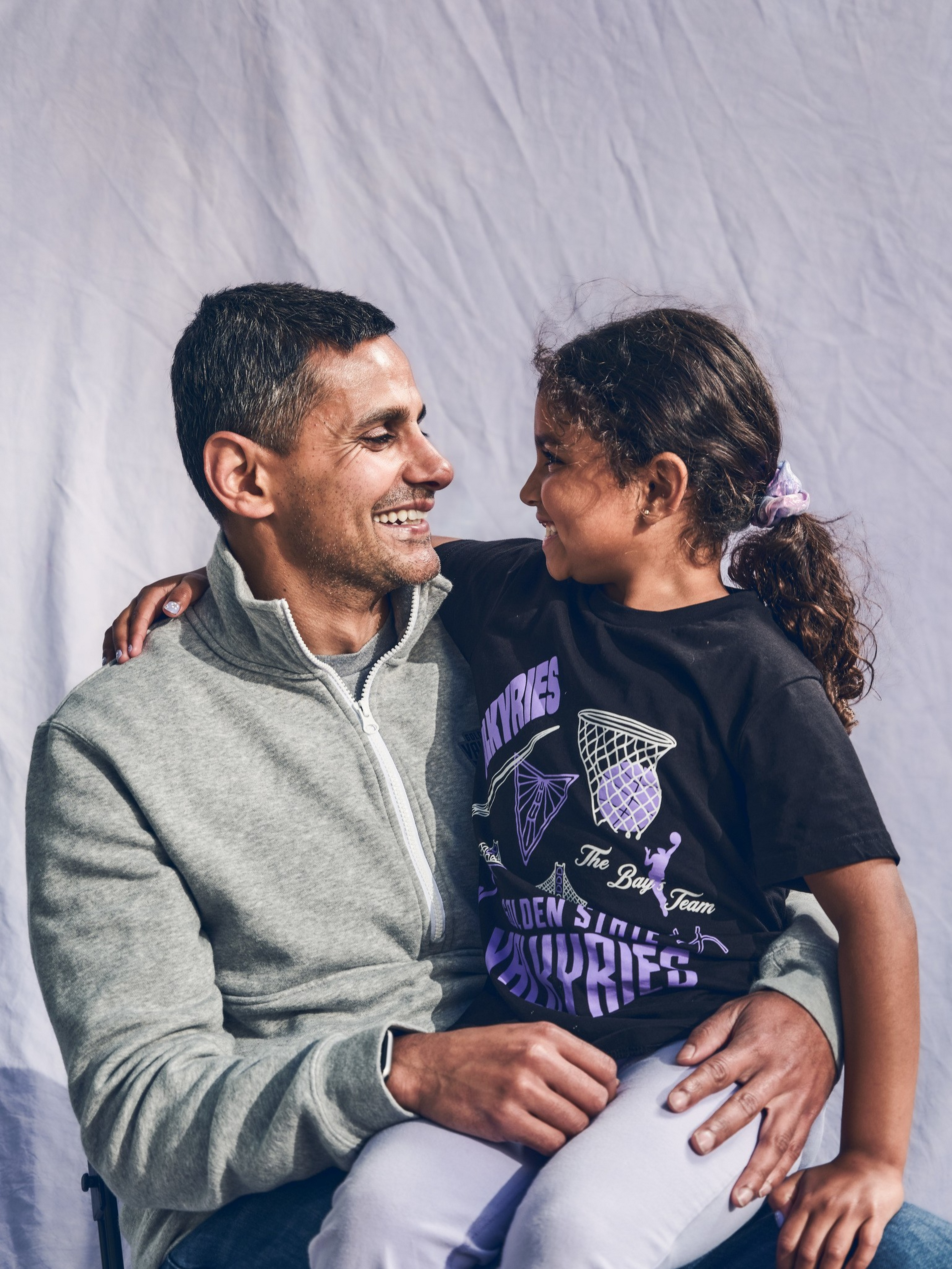 A man in a gray zip-up jacket smiles while holding a smiling girl in a black t-shirt and light pants on his lap, both looking at each other lovingly.