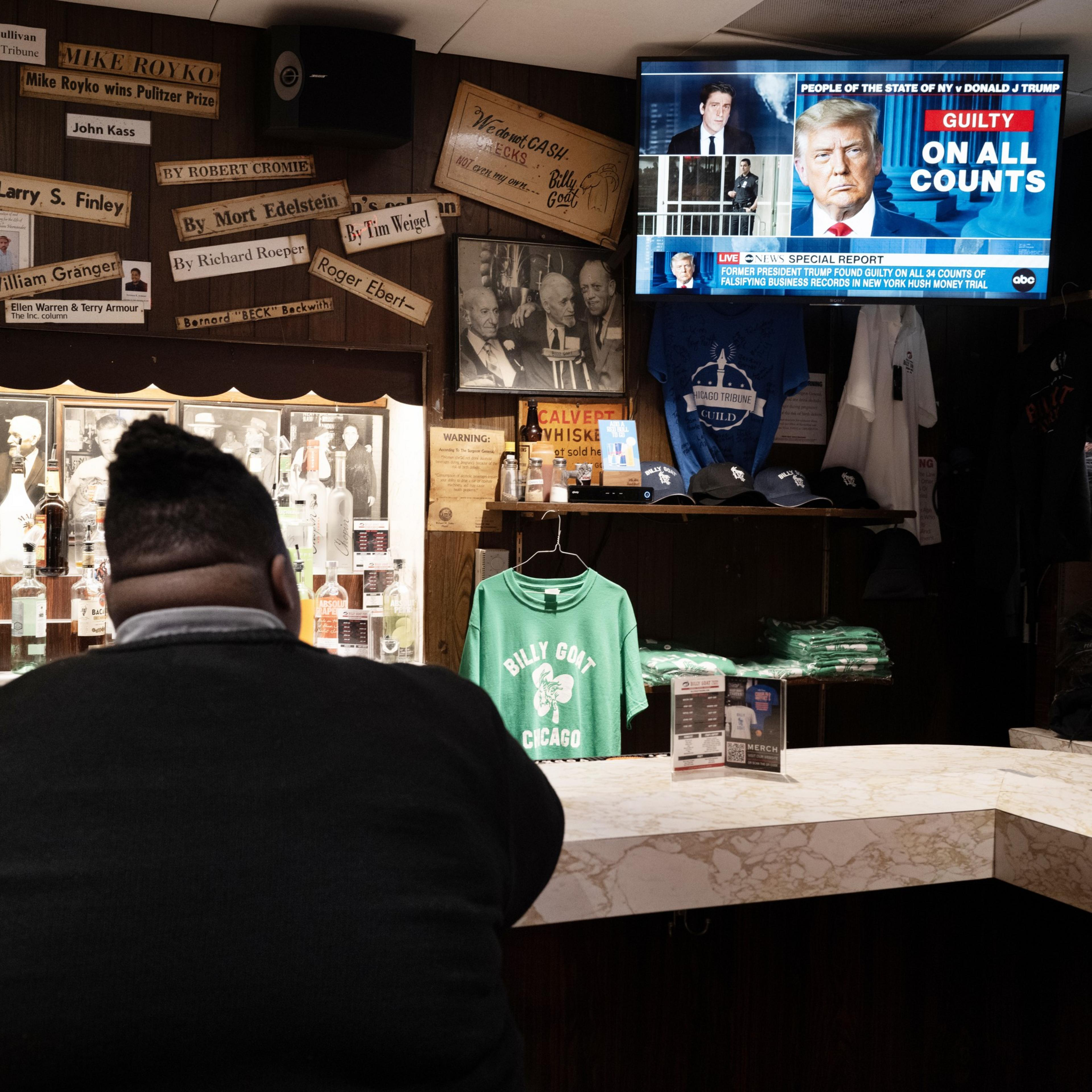 A person seated at a bar watches a TV showing news of Donald Trump found guilty on all counts. The bar has memorabilia and merchandise like hats and shirts.