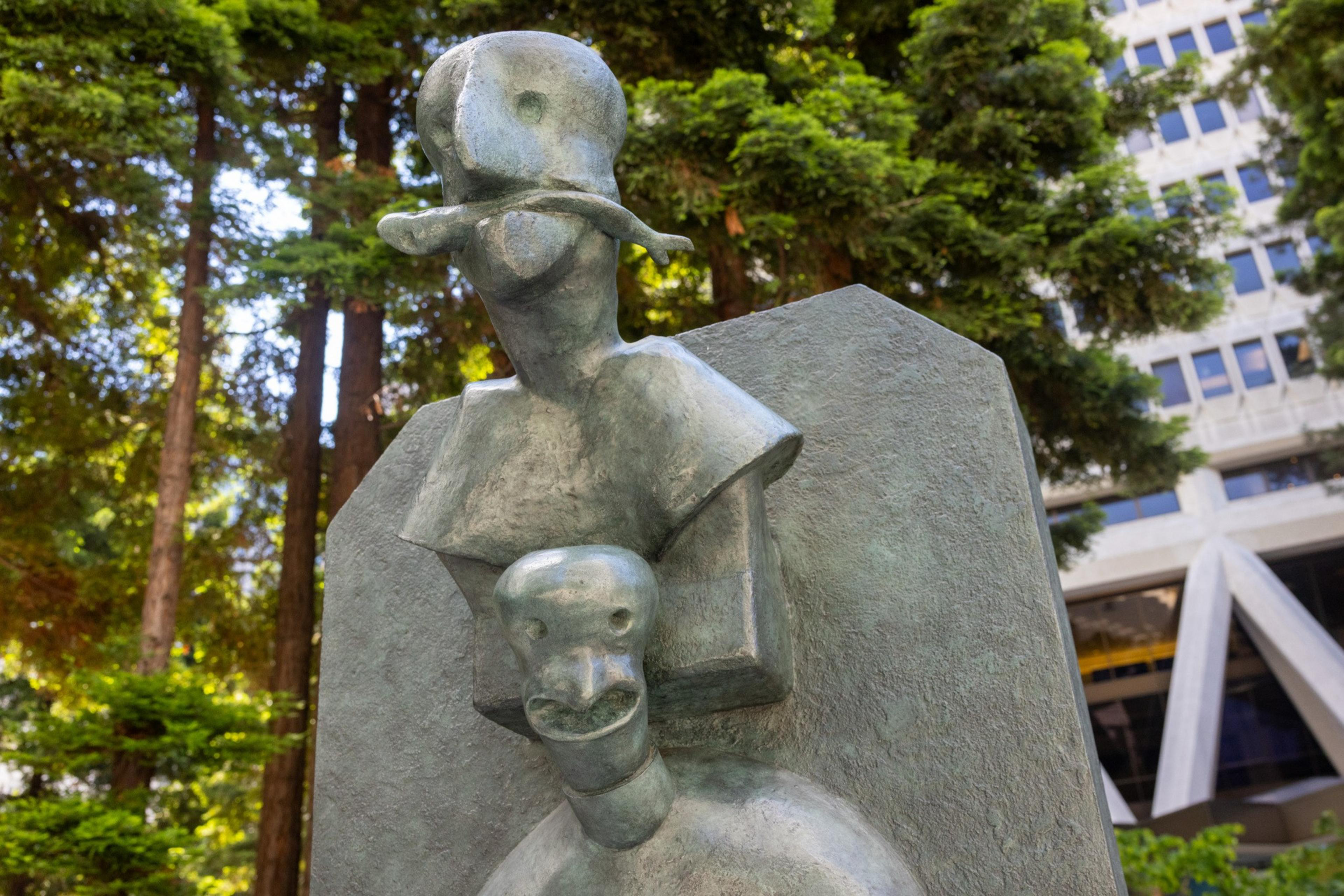 The image shows a metallic abstract sculpture of two stylized human figures with simplified features, set against a backdrop of tall trees and a modern building.