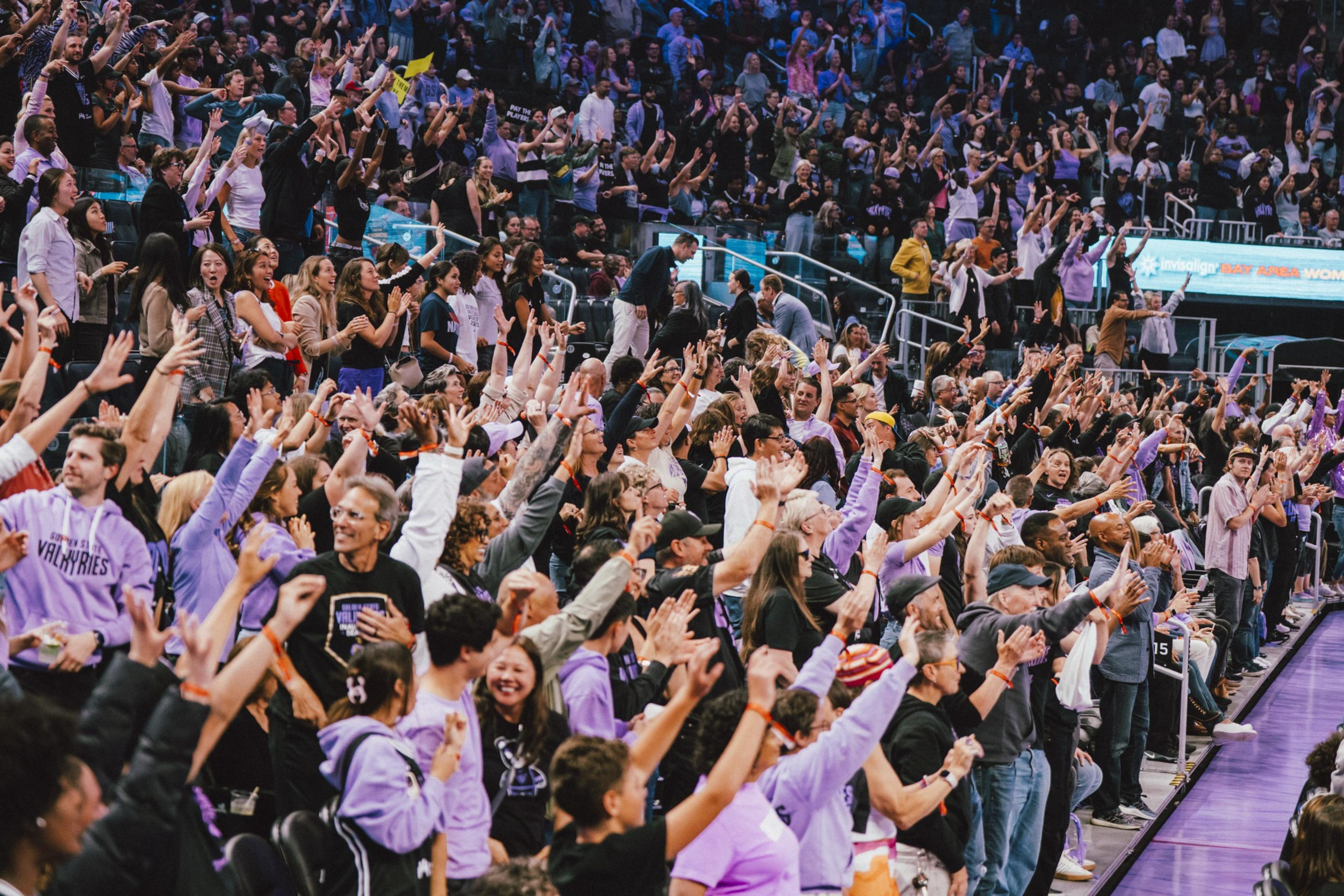 A large crowd of enthusiastic fans wearing purple and black raise their hands and cheer in an indoor sports arena.