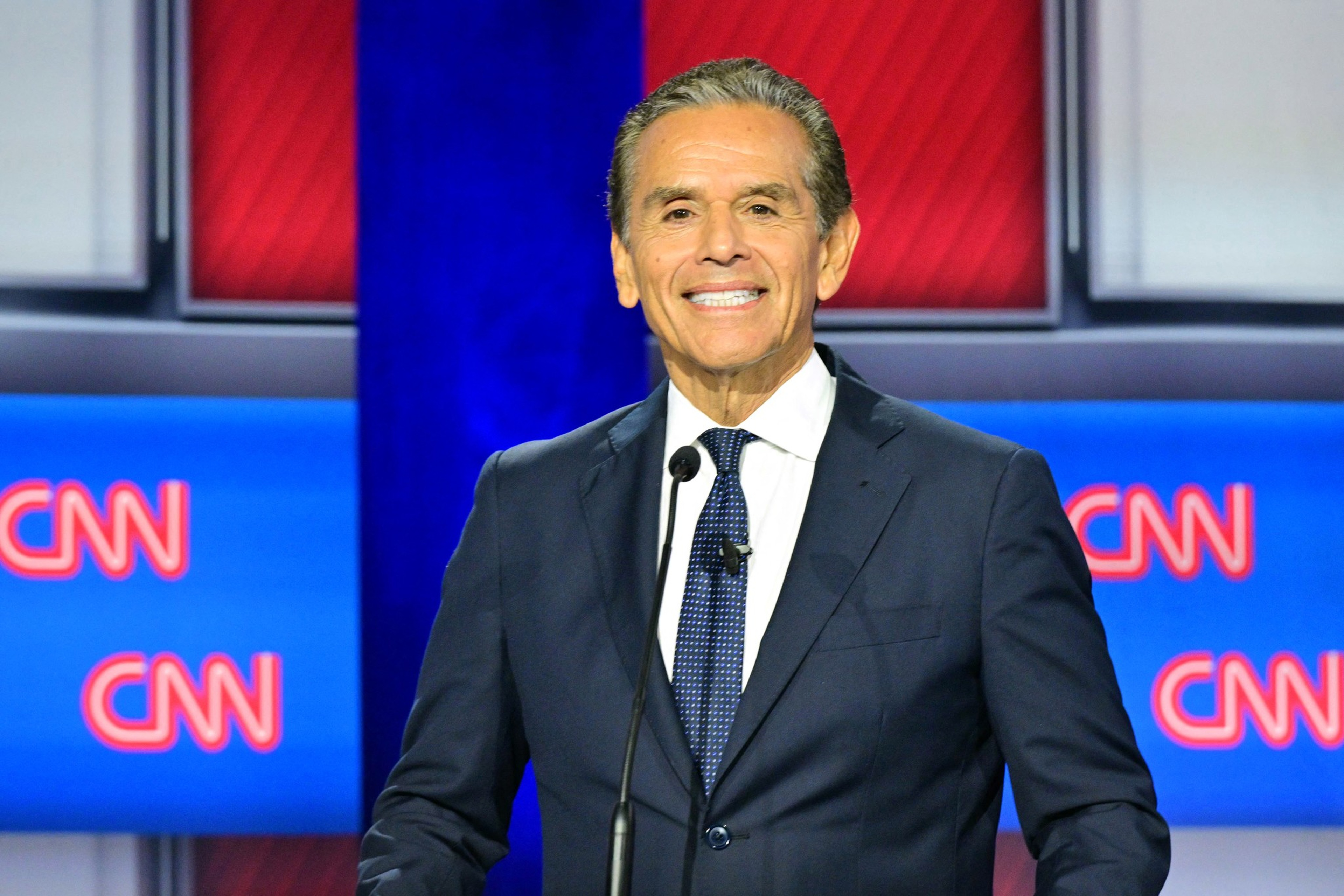 A man in a dark suit and tie smiles while standing behind a microphone with CNN logos on screens behind him.