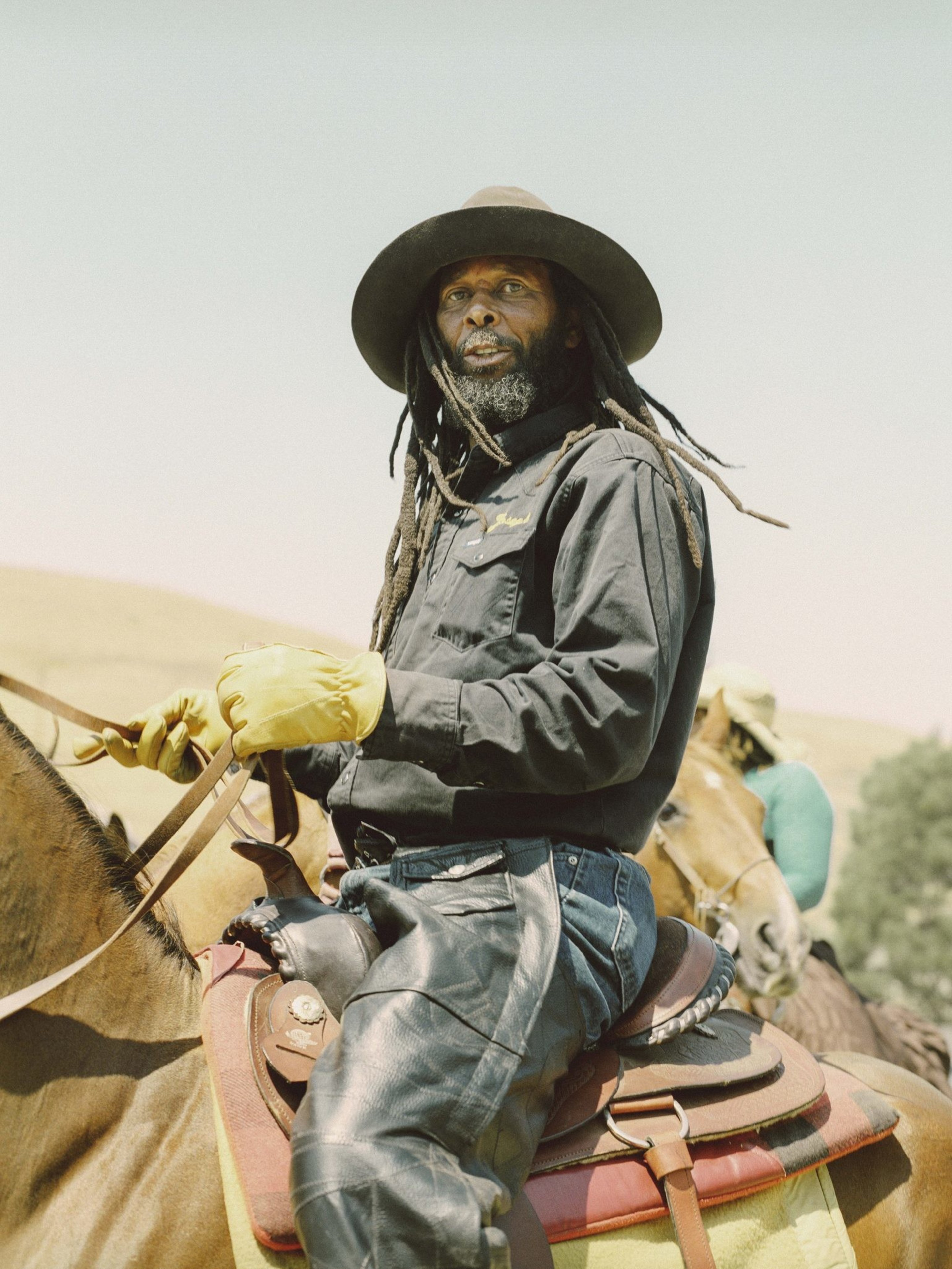 A person with dreadlocks and a wide-brimmed hat rides a horse. They wear a black jacket, leather chaps, and yellow gloves, with a sunny landscape in the background.