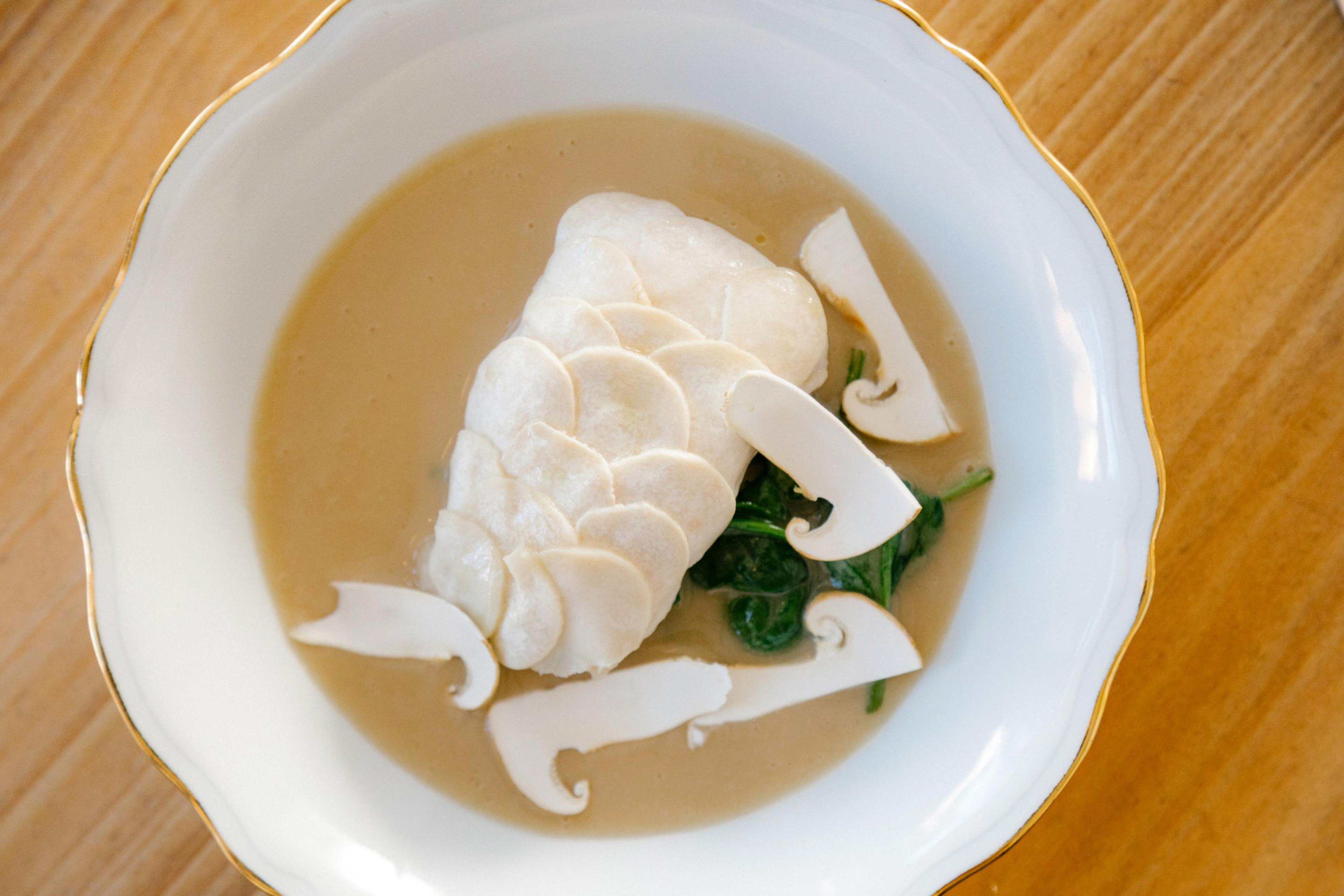 A plated dish with a white fish fillet topped with thin slices, served on sautéed greens and creamy mushroom sauce, garnished with sliced raw mushrooms.