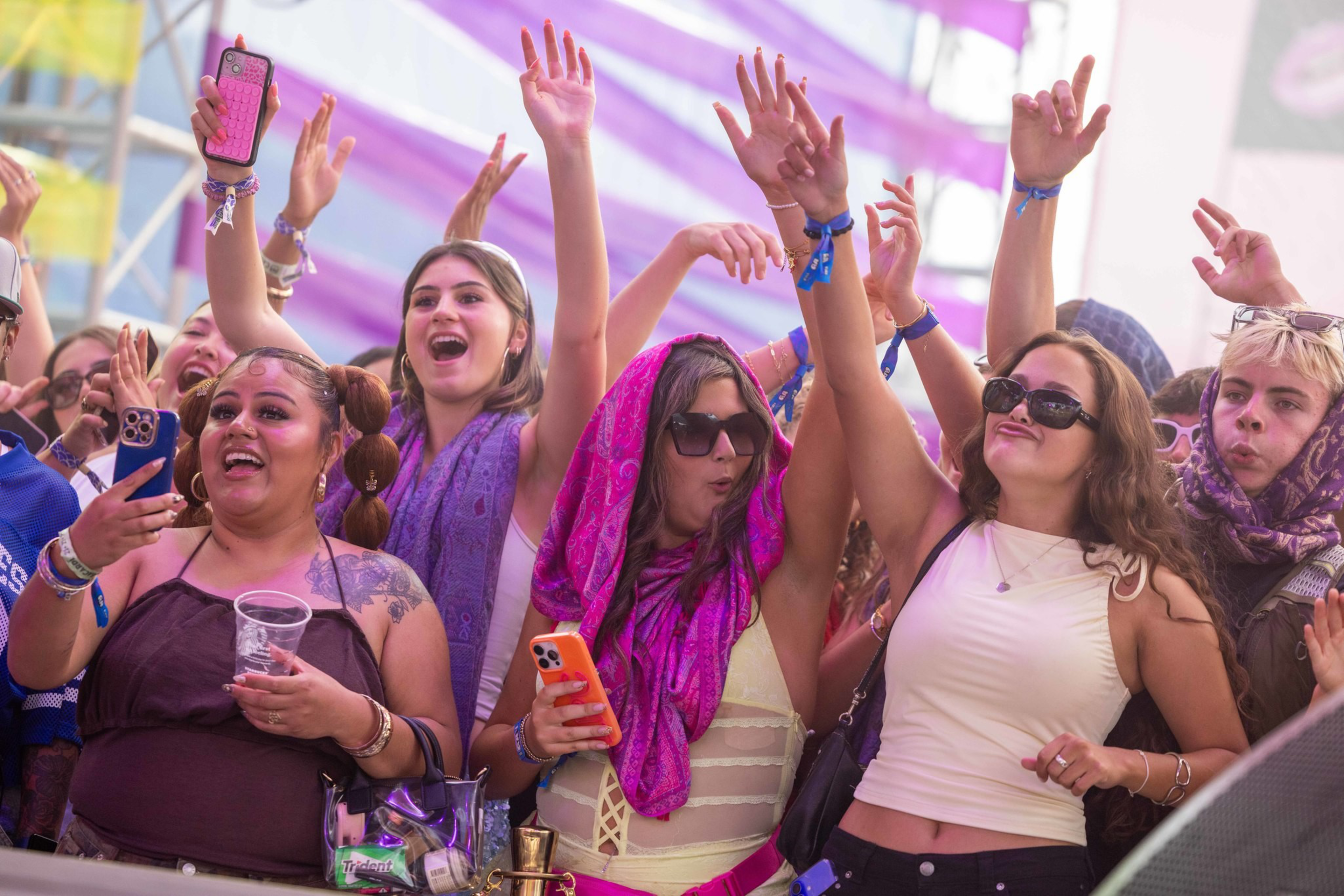 A group of young people at a colorful event cheer with hands raised, some holding phones, dressed in casual and vibrant attire under a striped purple canopy.