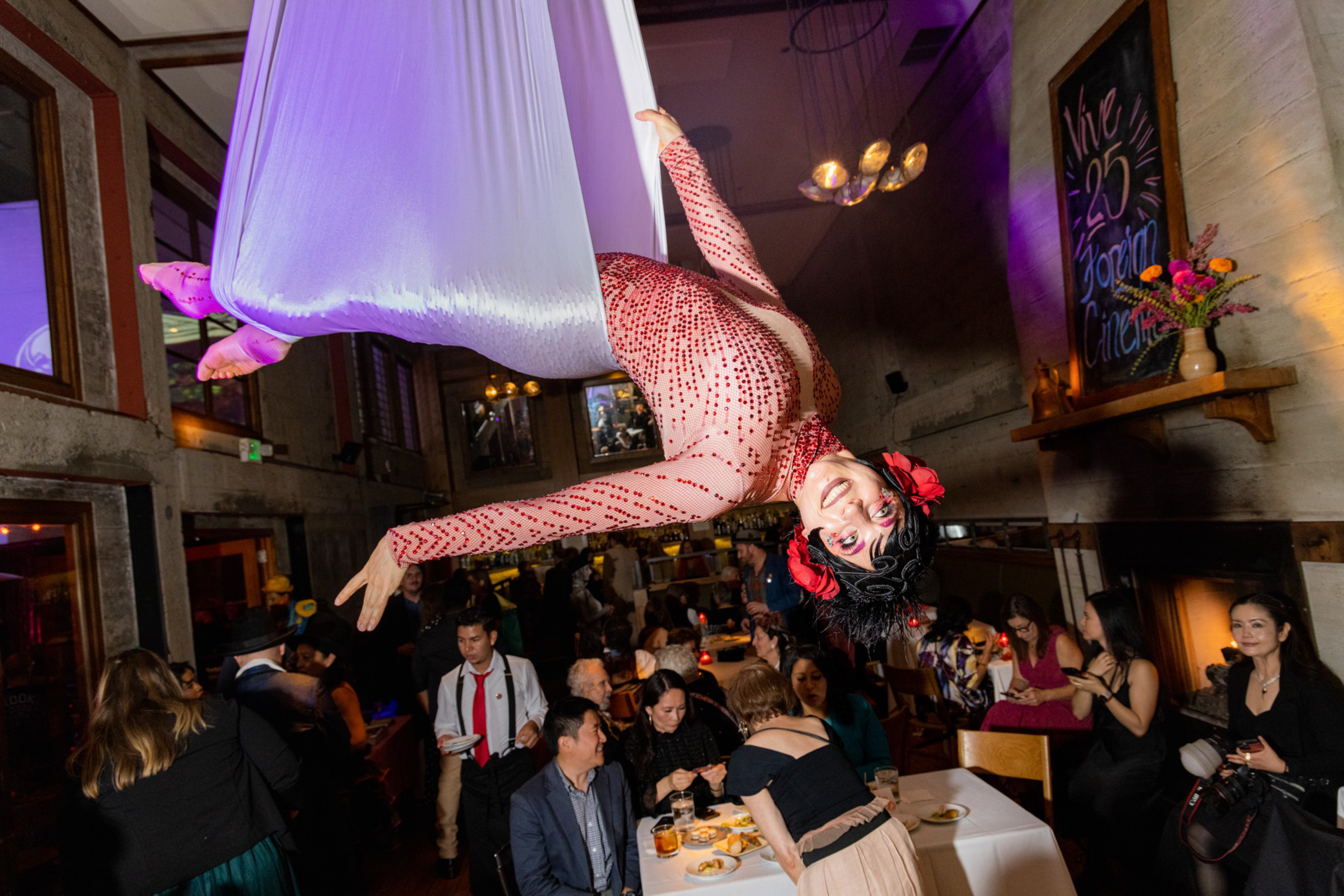 An aerial performer in a red, sequined bodysuit and pink shoes hangs from purple fabric above a lively gathering of people in a warmly lit venue.