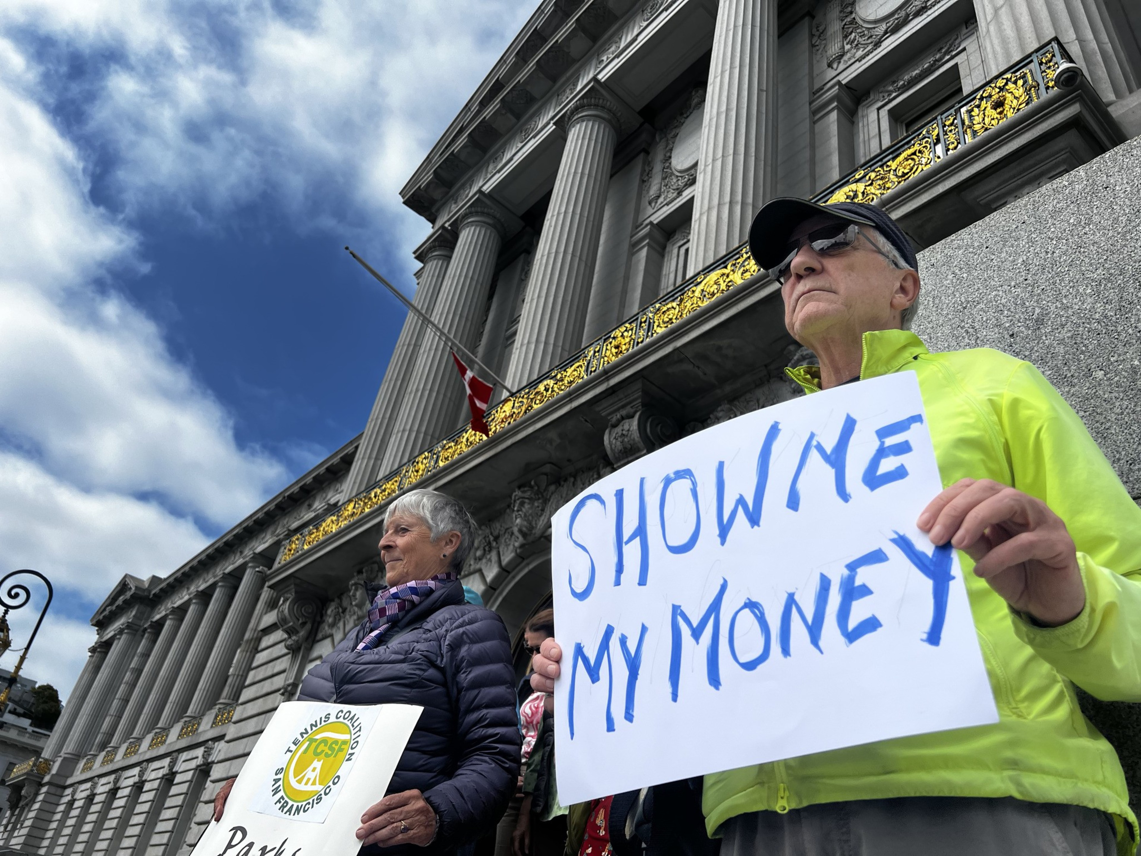 Two people stand in front of a grand building with columns. One holds a sign reading &quot;SHOW ME MY MONEY,&quot; while the other holds a &quot;Tennis Coalition San Francisco&quot; sign.
