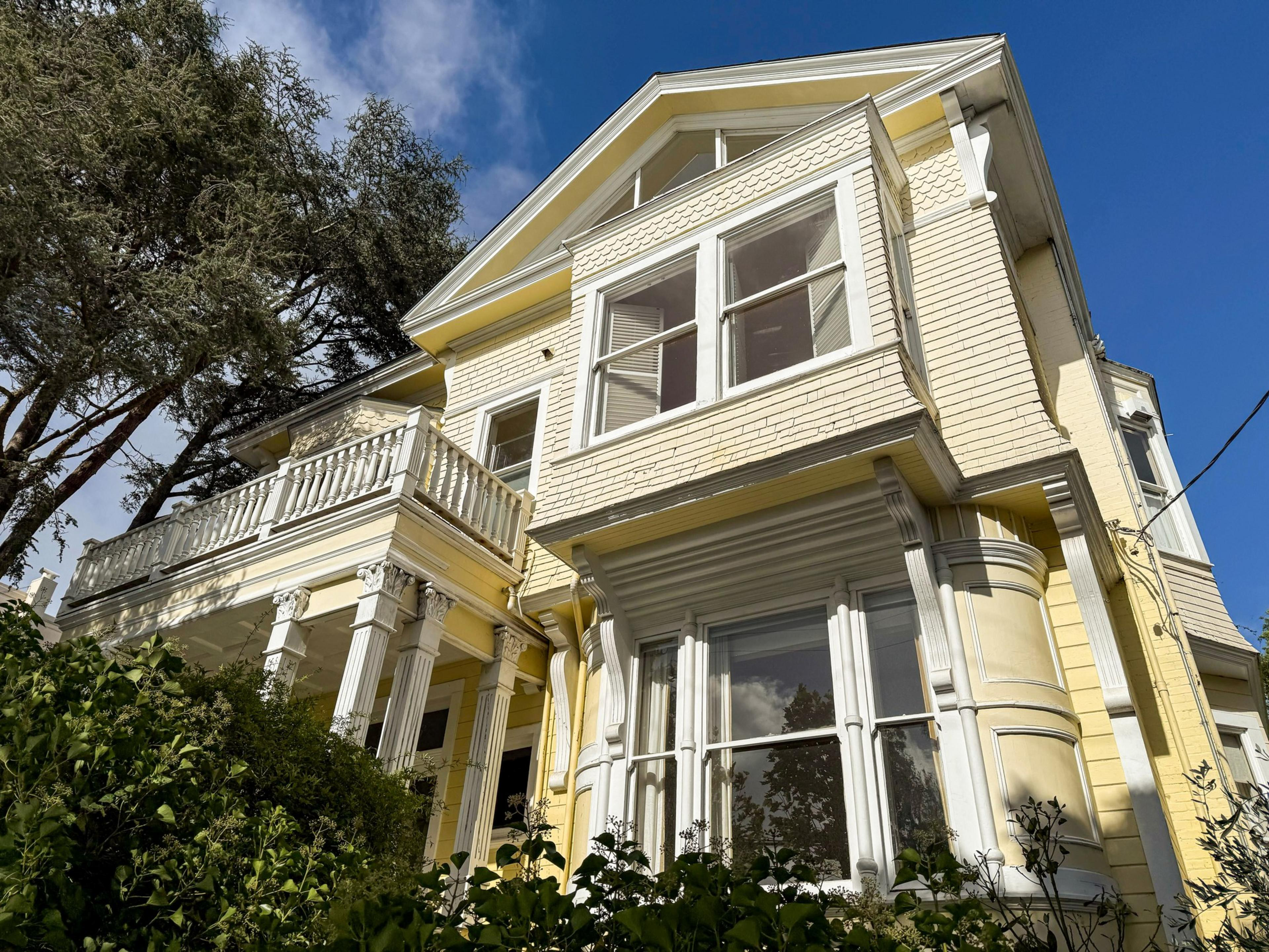 A yellow Victorian-style house with white trim, large windows, a balcony with columns, and green bushes in front under a clear blue sky.
