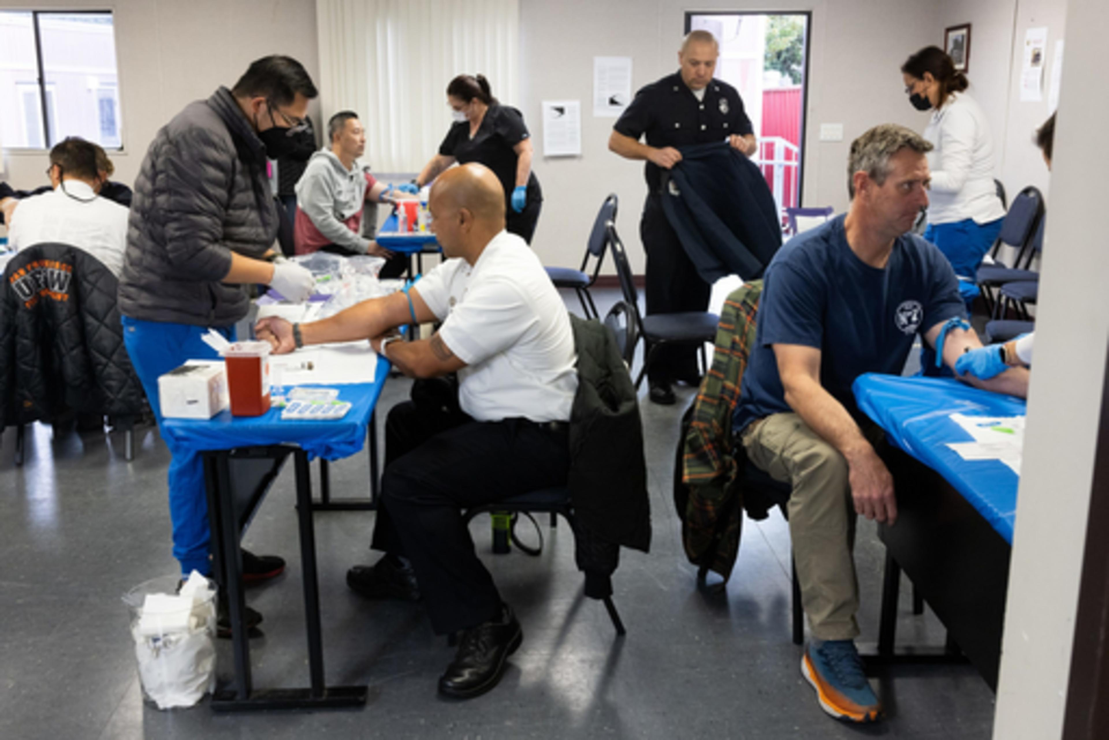 Several men are seated at tables getting blood drawn by healthcare workers in a clinic room, with one officer standing and others waiting nearby.
