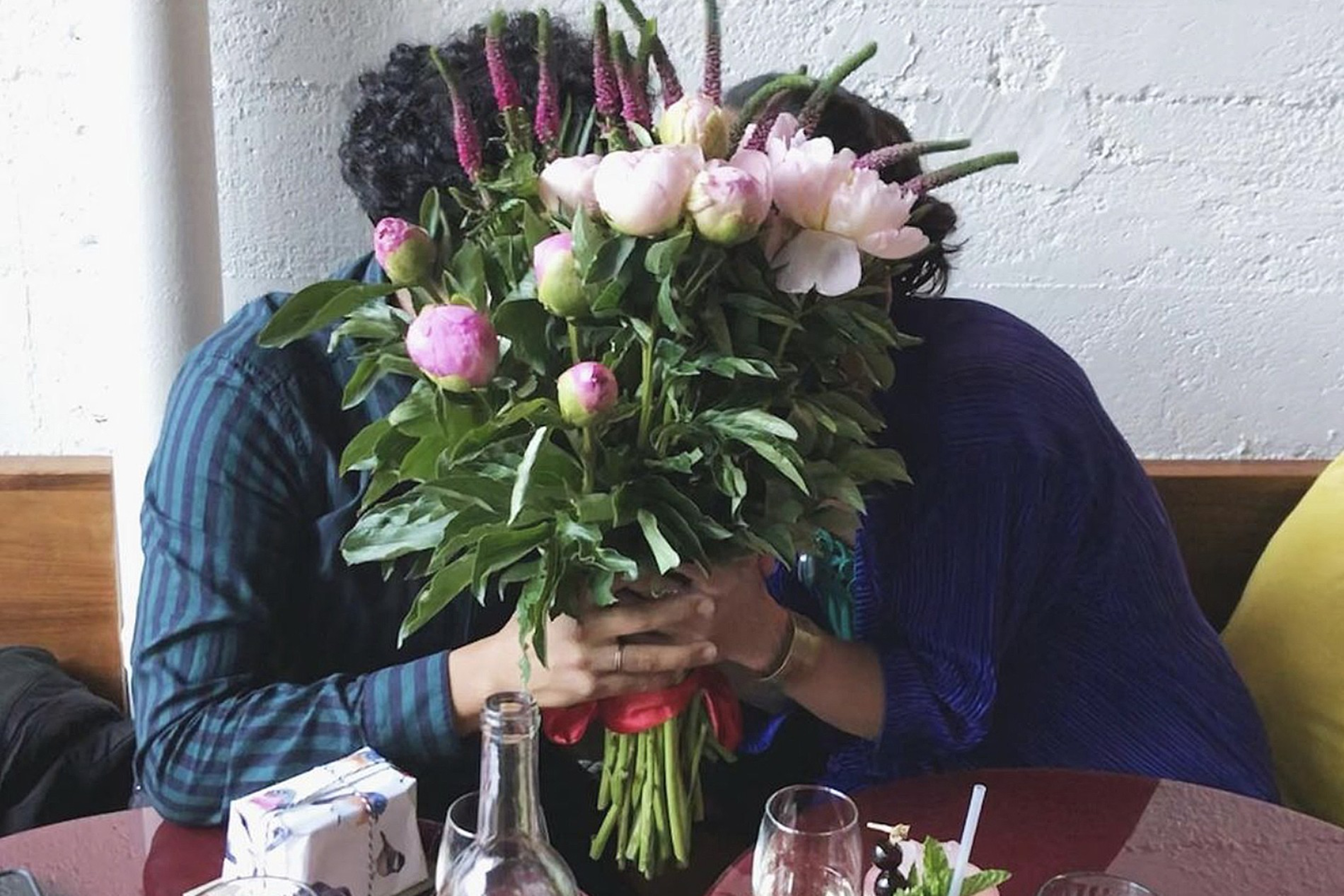Two people sit closely at a table, their faces hidden behind a large bouquet of pink and purple flowers with green leaves.
