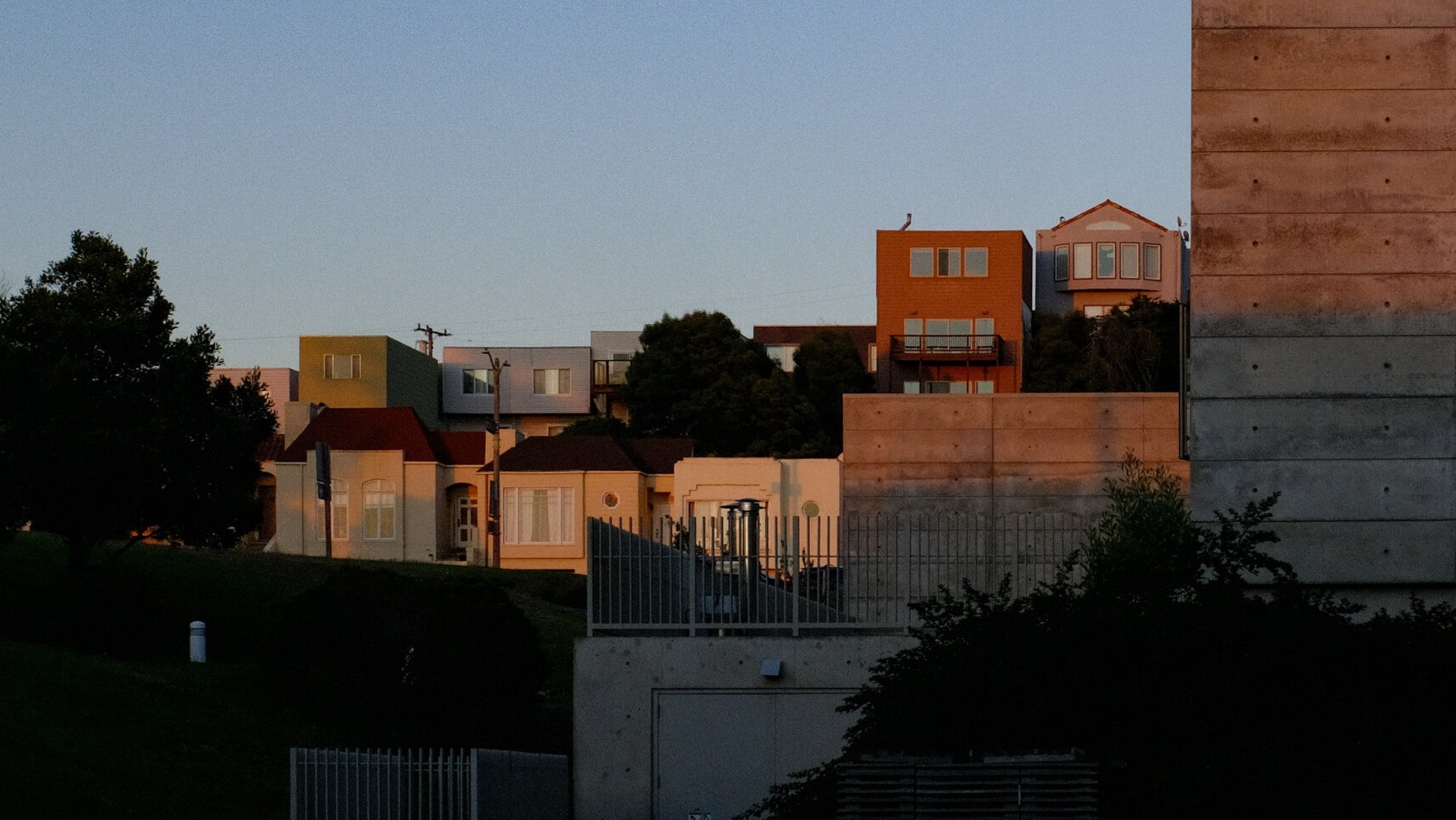 Colorful houses sit atop a hill in the warm light of sunset, with large concrete walls and trees in the foreground.