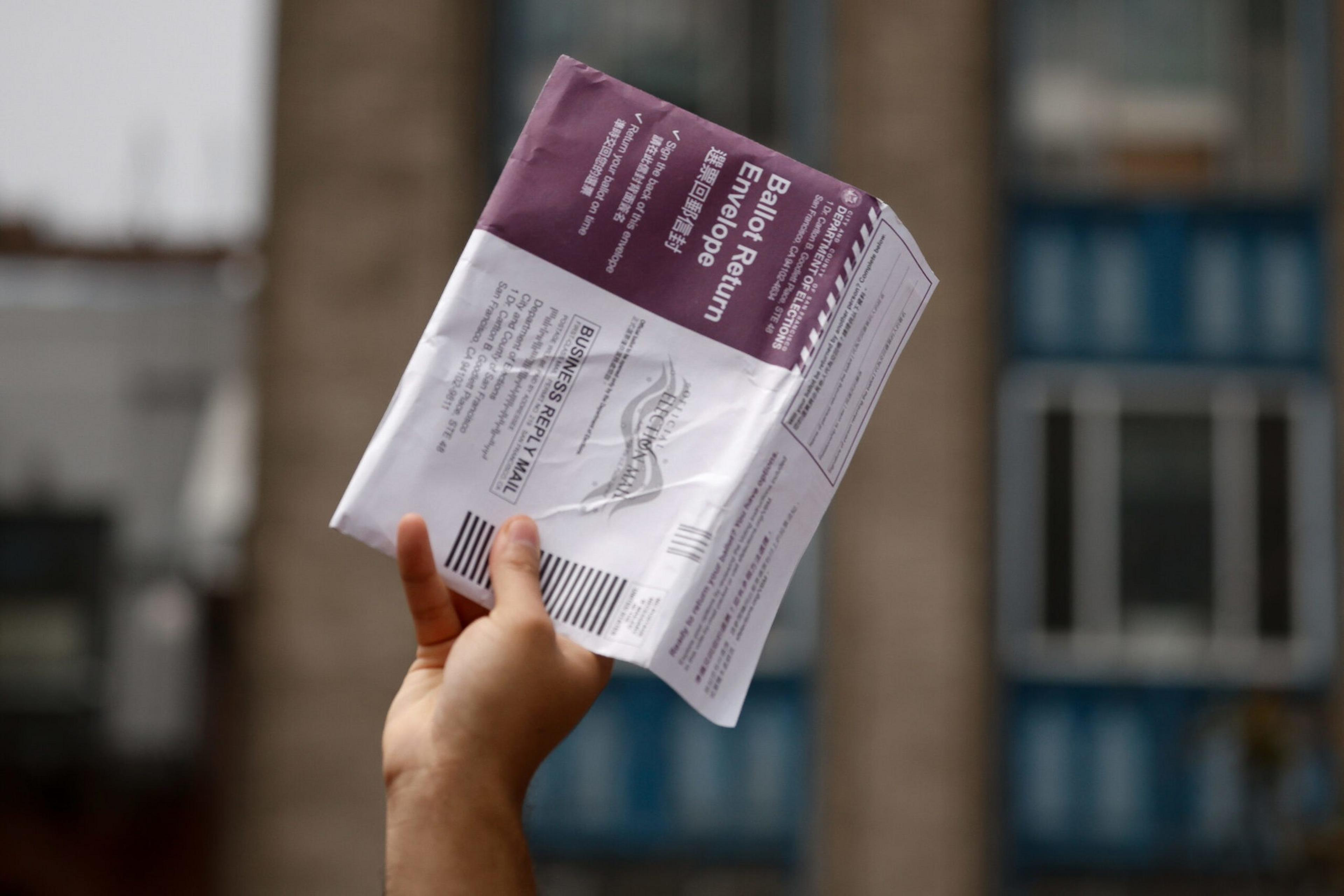 A hand holds up a ballot return envelope against a blurred urban background with blue and beige buildings.