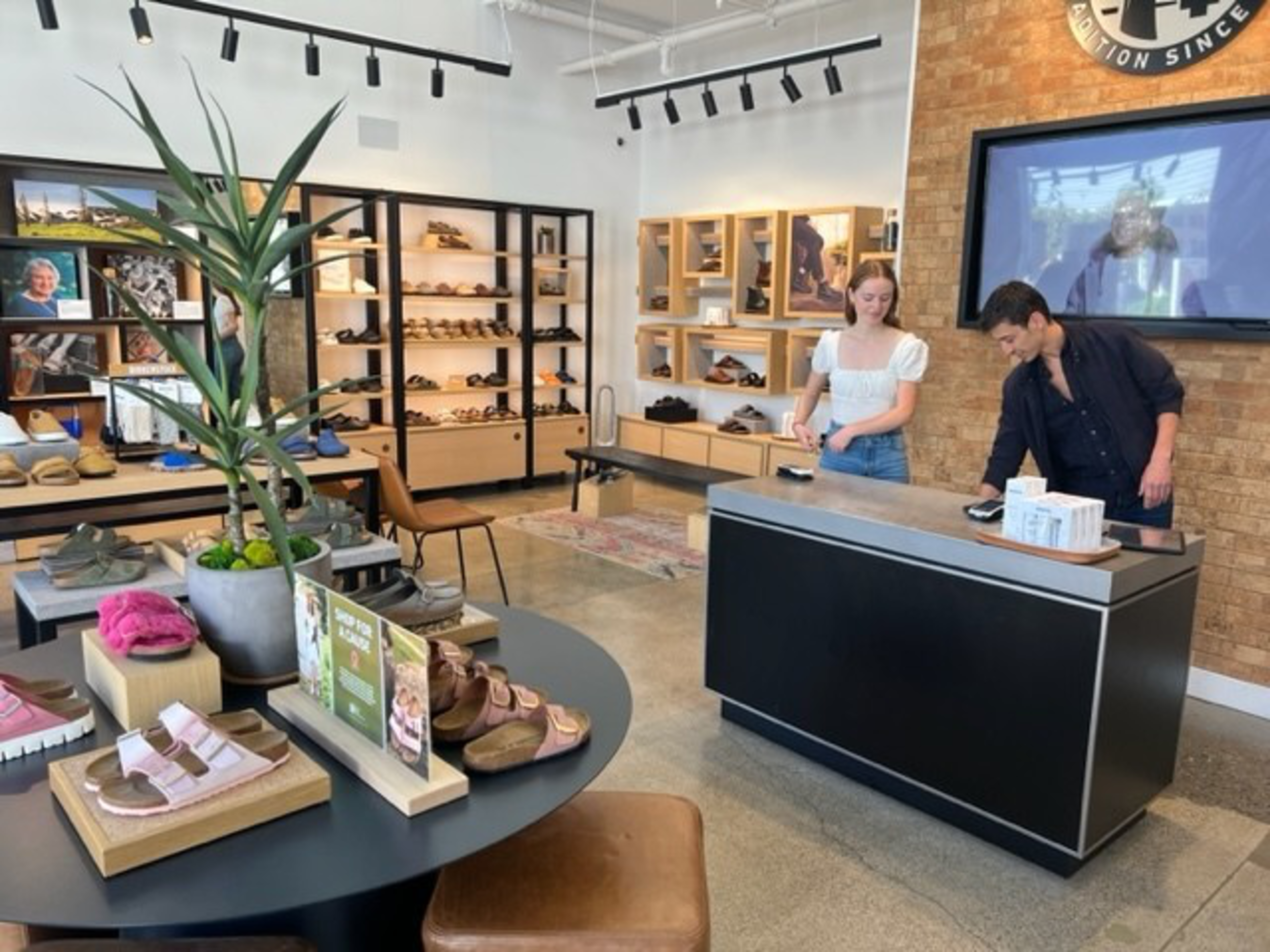 A shoe store features modern displays with sandals and slippers, a large potted plant on a table, and two people at the checkout counter.