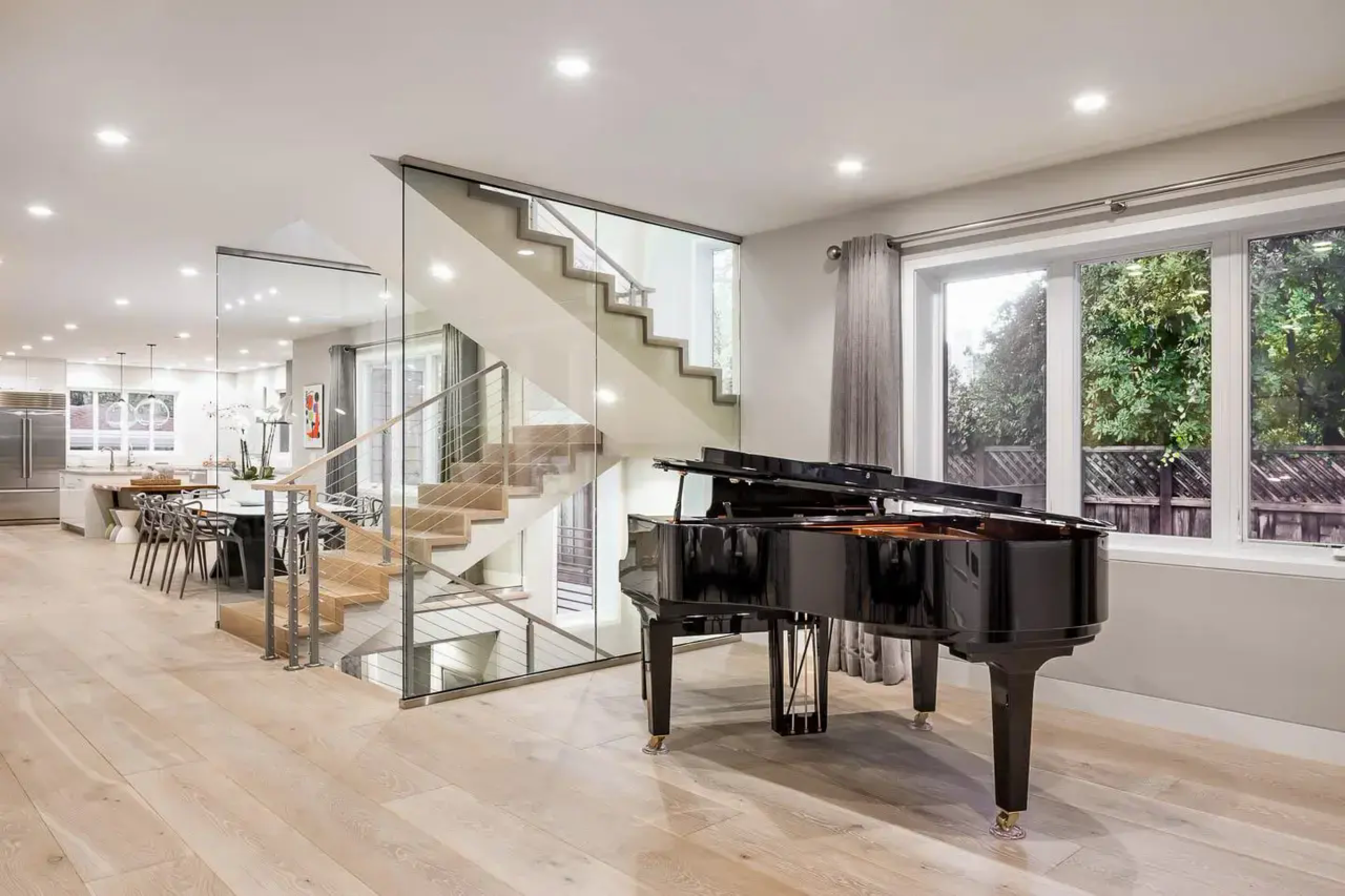 A black grand piano sits on light wood flooring beside large windows with gray curtains, near a modern glass-encased staircase and a bright open kitchen.