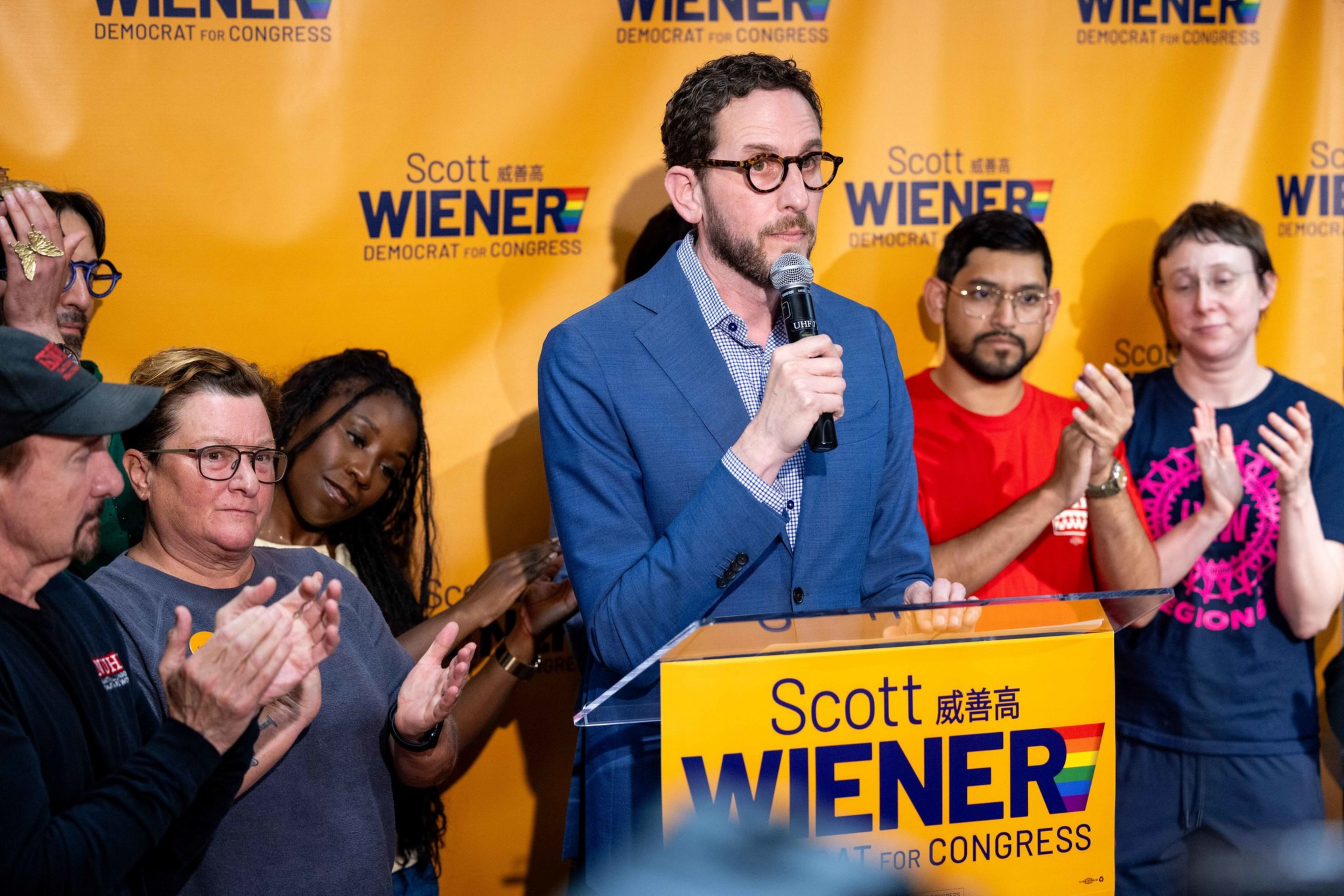 A man in a blue suit speaks into a microphone at a podium with “Scott Wiener Democrat for Congress” signs, while a diverse group behind him applauds.