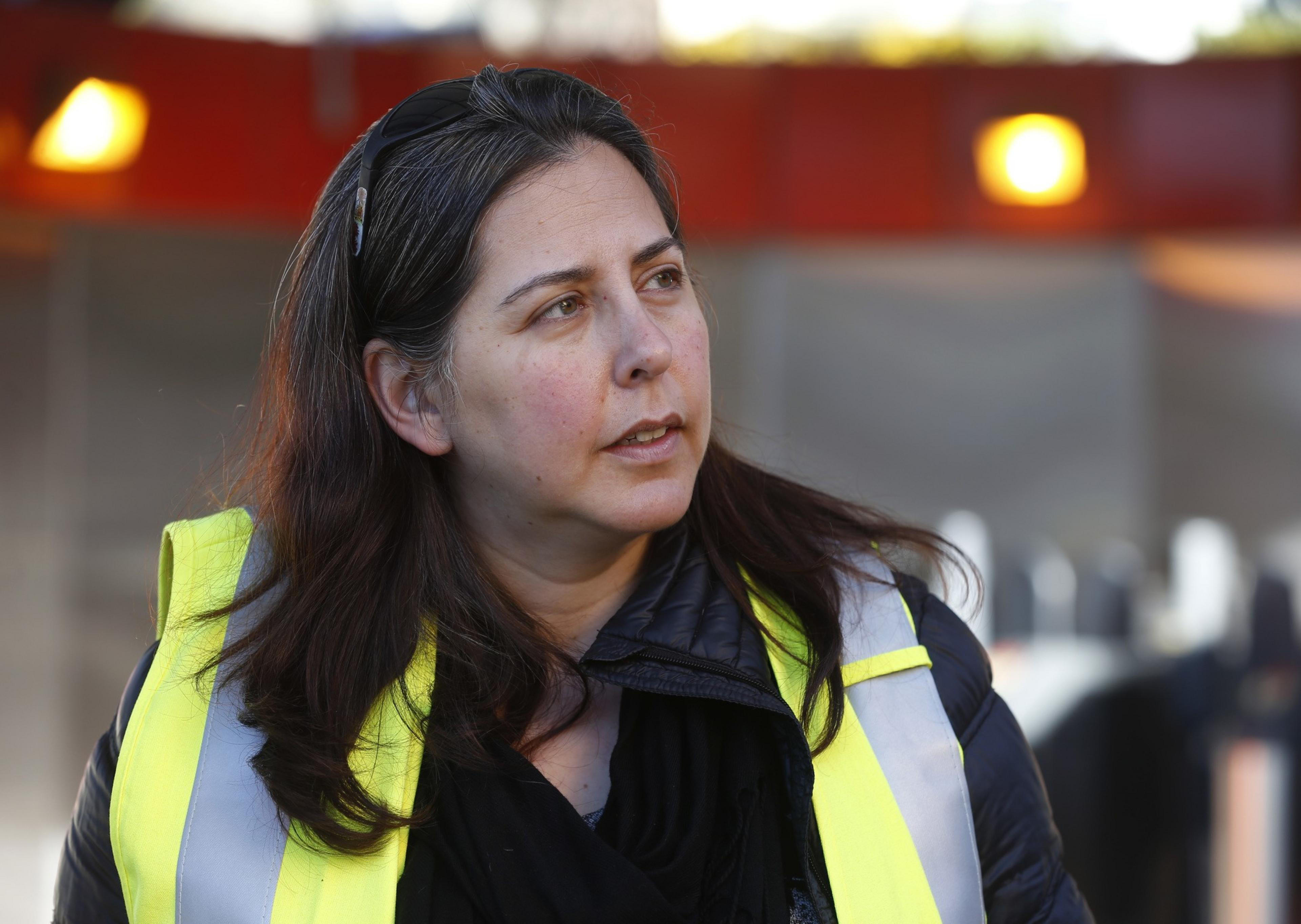 A woman with long, dark hair wears a reflective safety vest and a black jacket. She looks to the side, with a blurred background featuring bright lights.