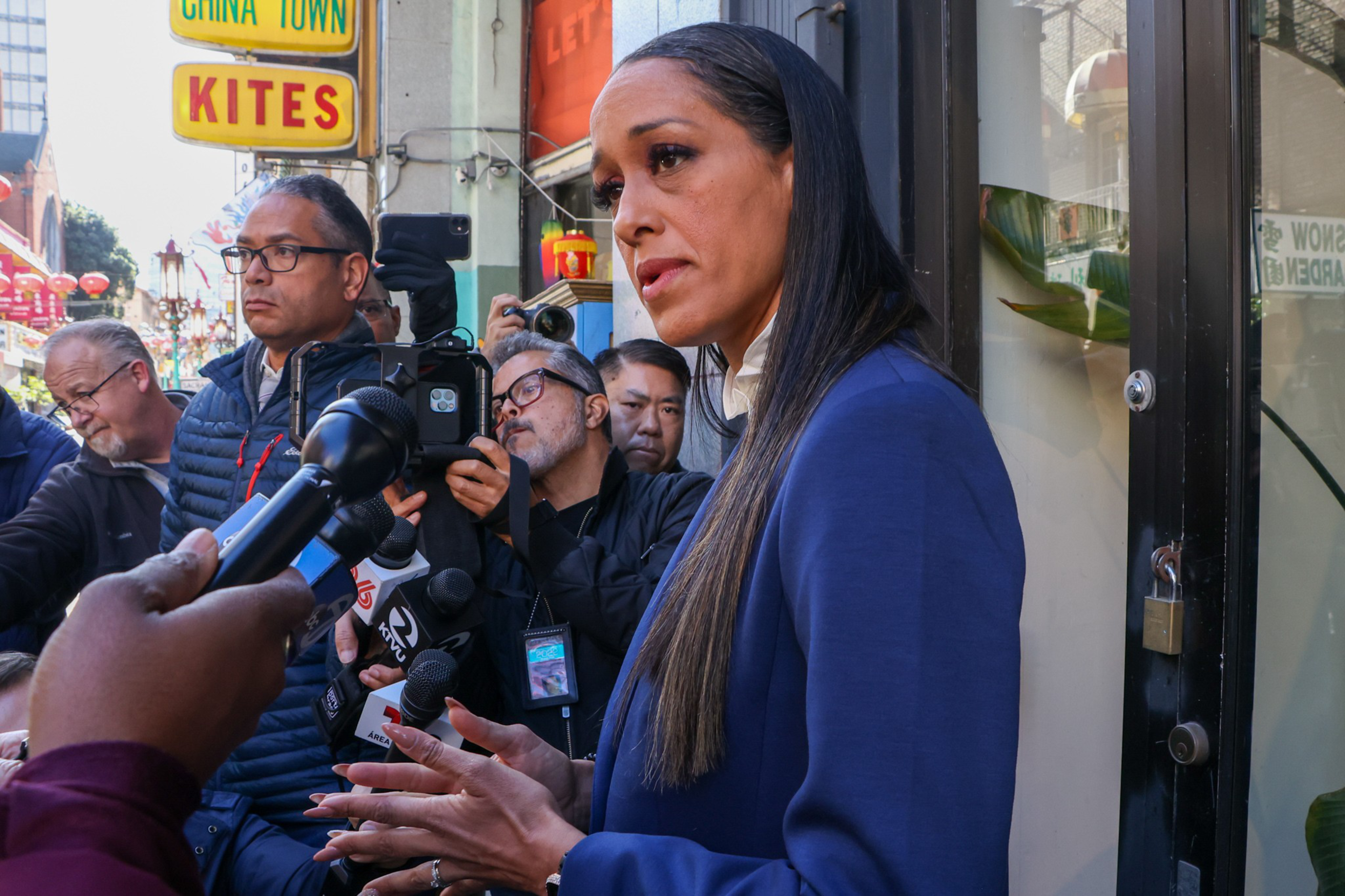 A woman in a blue blazer speaks to reporters holding microphones and cameras in an outdoor urban setting near a "CHINA TOWN KITES" sign.