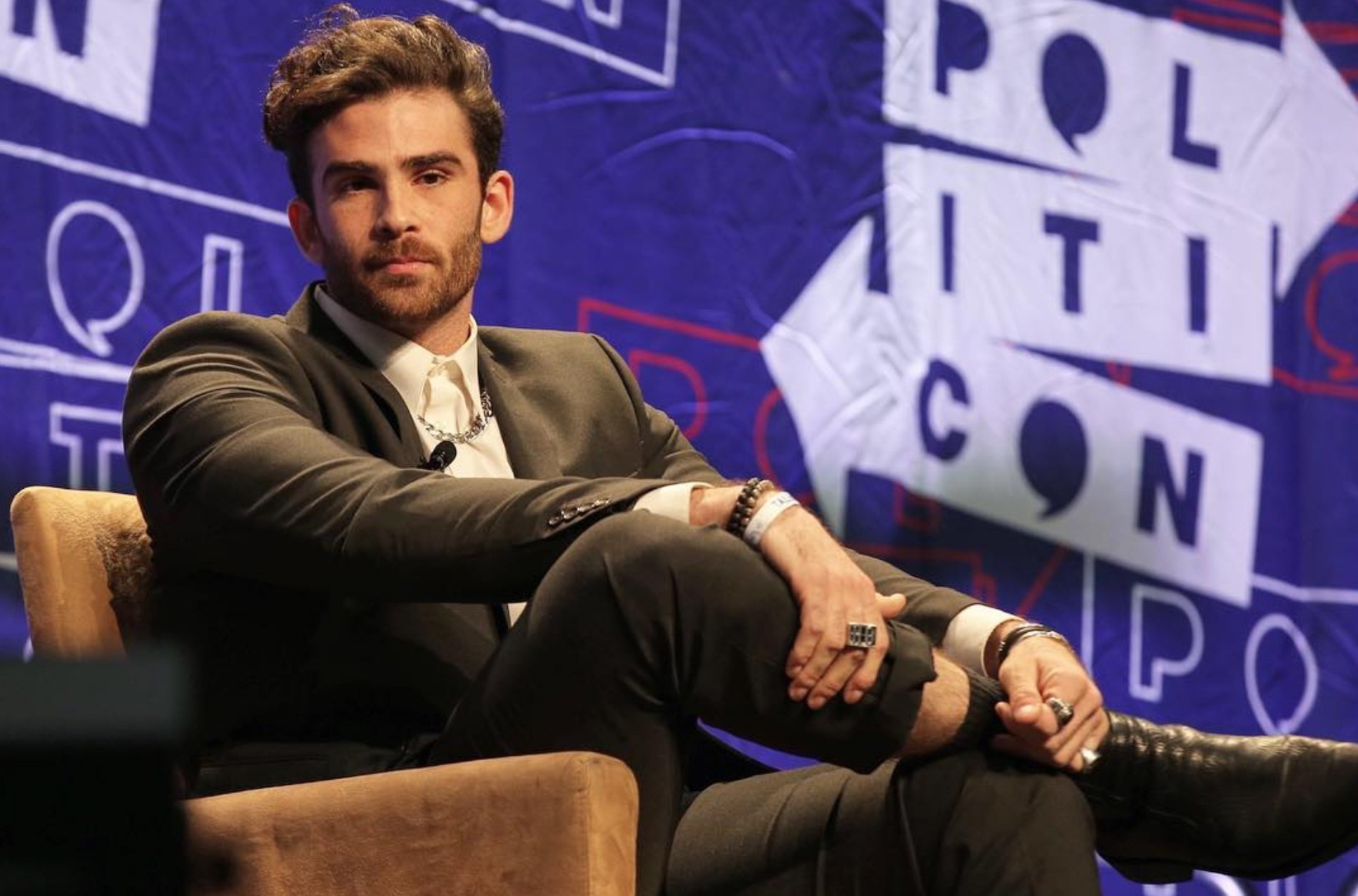 A man with styled hair and a beard sits cross-legged on a chair, wearing a dark suit and white shirt, in front of a purple backdrop with “POLITICON” text.
