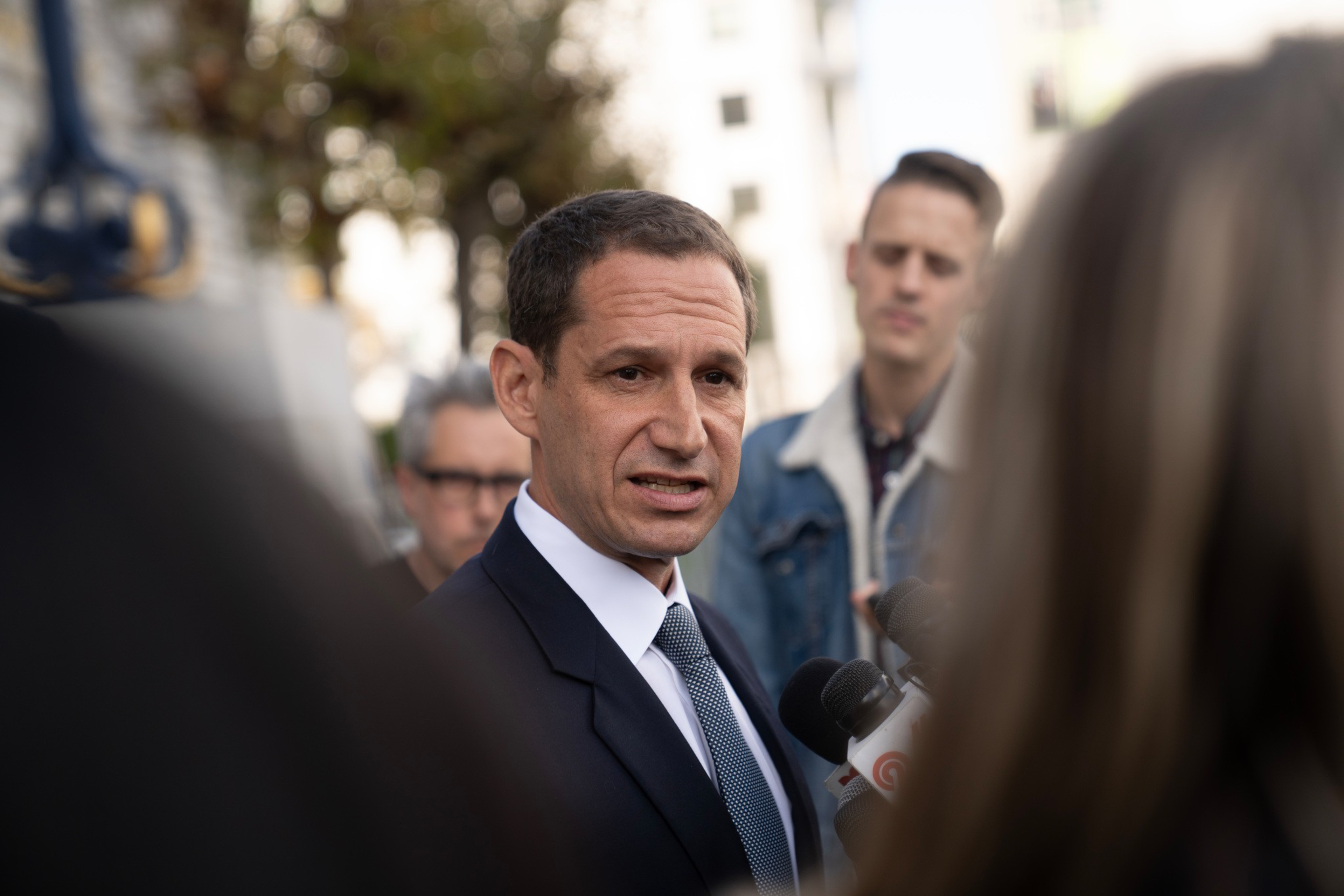 A man in a suit and tie speaks seriously to reporters holding microphones, while two people listen attentively in the blurred background.