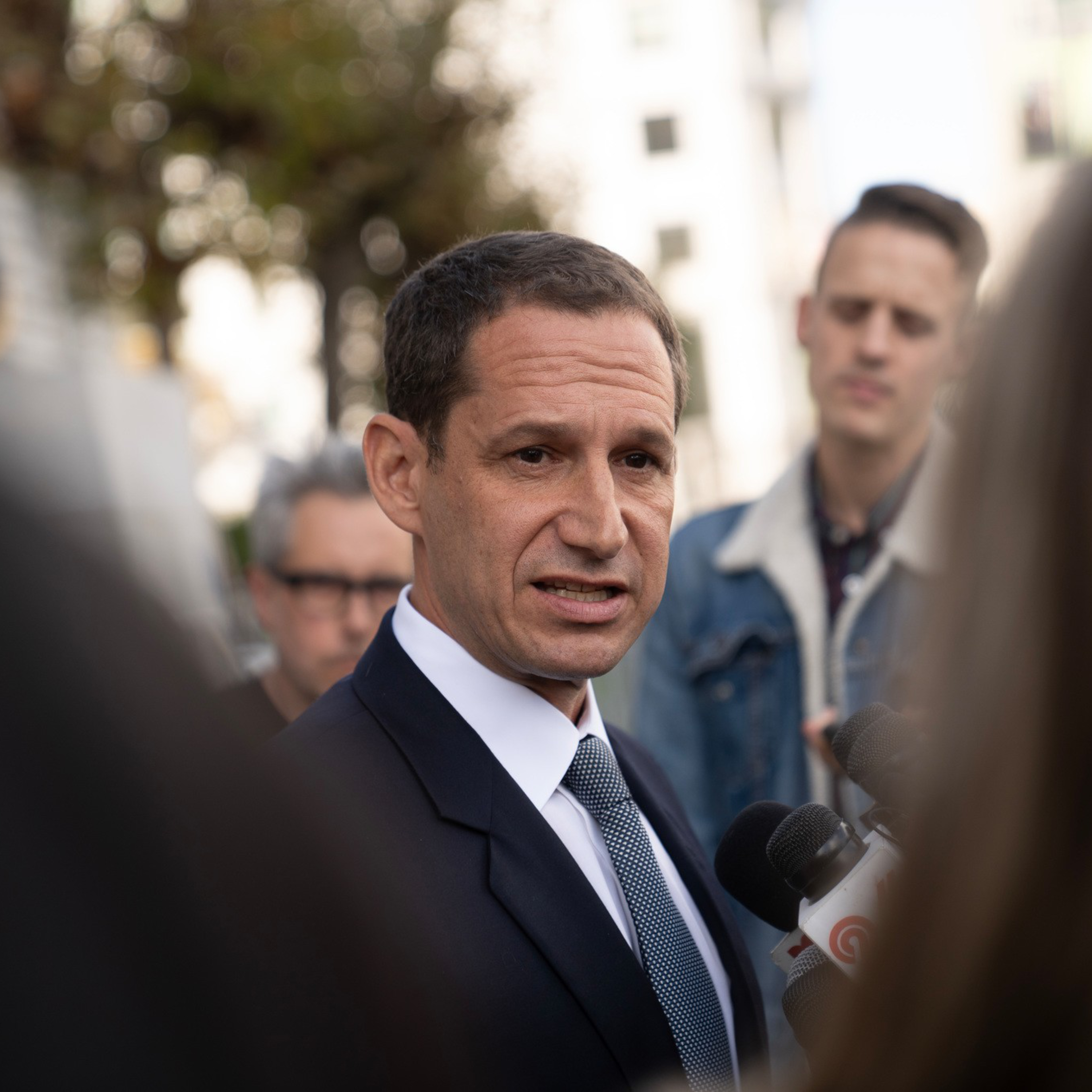 A man in a suit and tie speaks seriously to reporters holding microphones, while two people listen attentively in the blurred background.