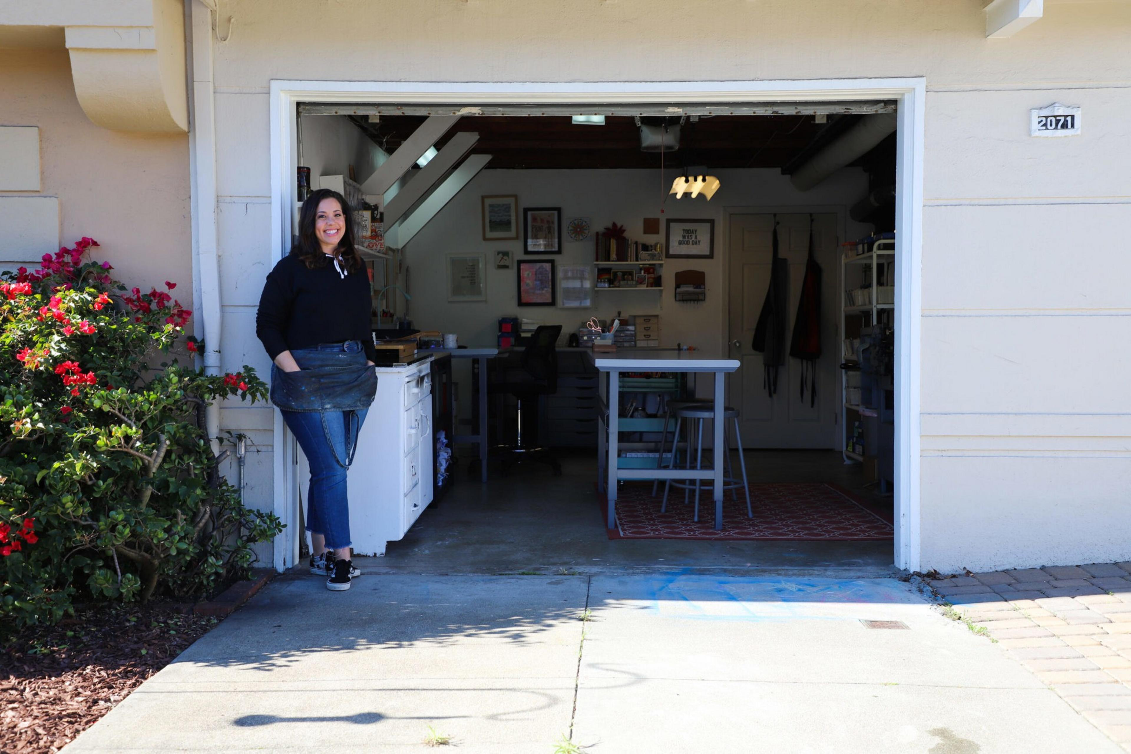 A woman stands smiling at the open garage door of a workshop with a table, stools, art supplies, and framed pictures on the walls inside.