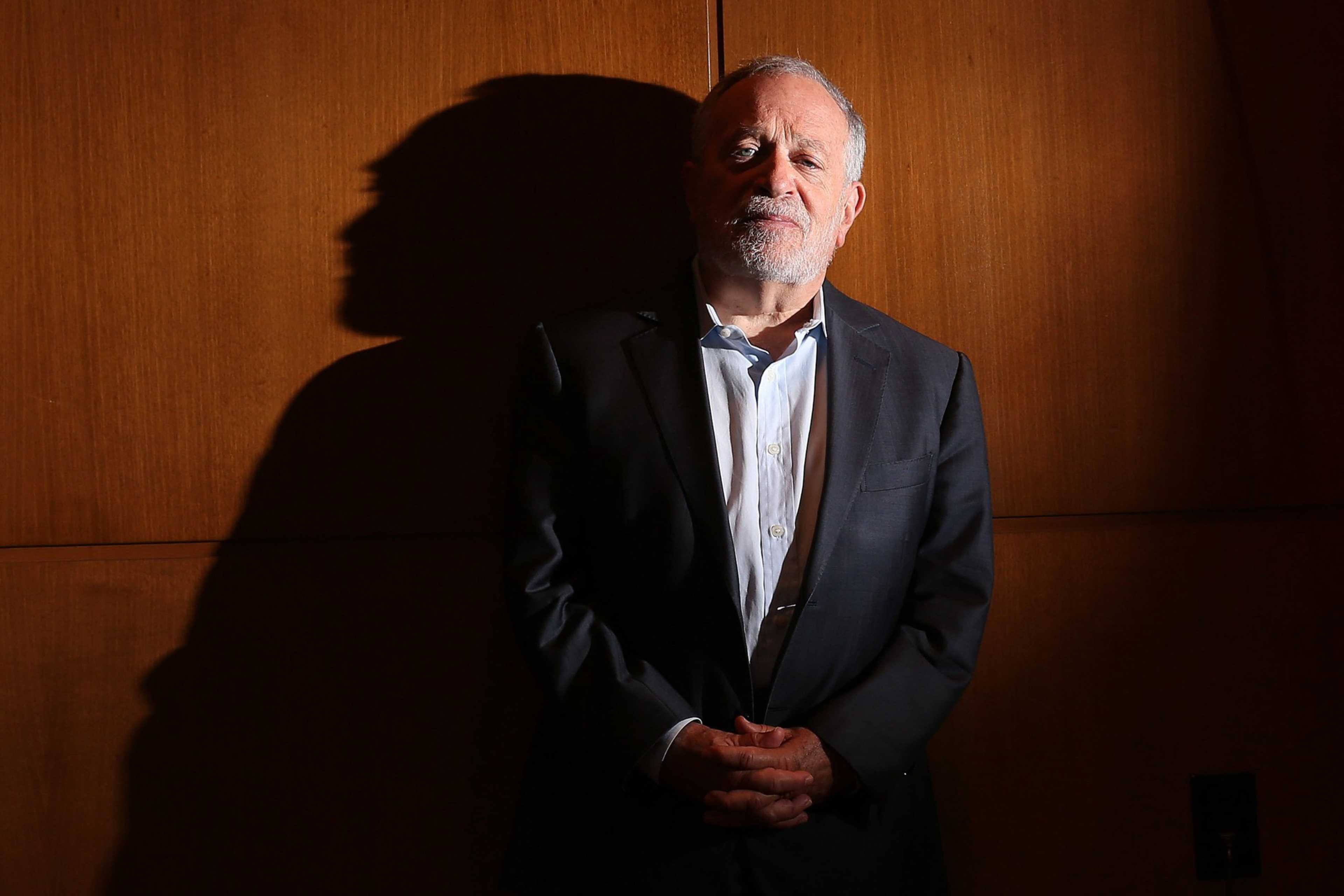 An older man with gray hair and beard stands with hands clasped, wearing a dark suit and white shirt, casting a large shadow on a wooden wall.