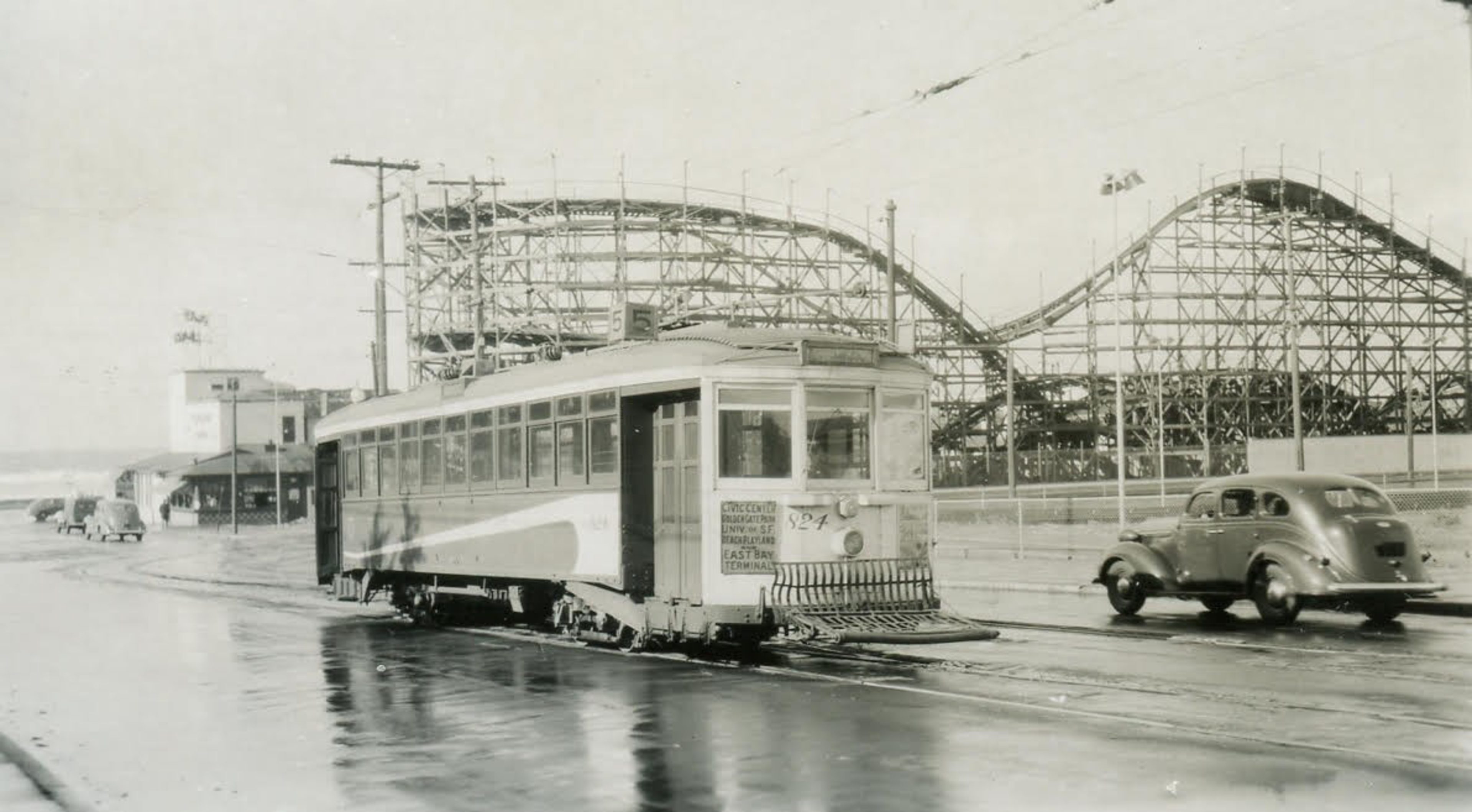 A streetcar on wet tracks runs past a wooden roller coaster, with a vintage car driving nearby on a rain-slicked road.