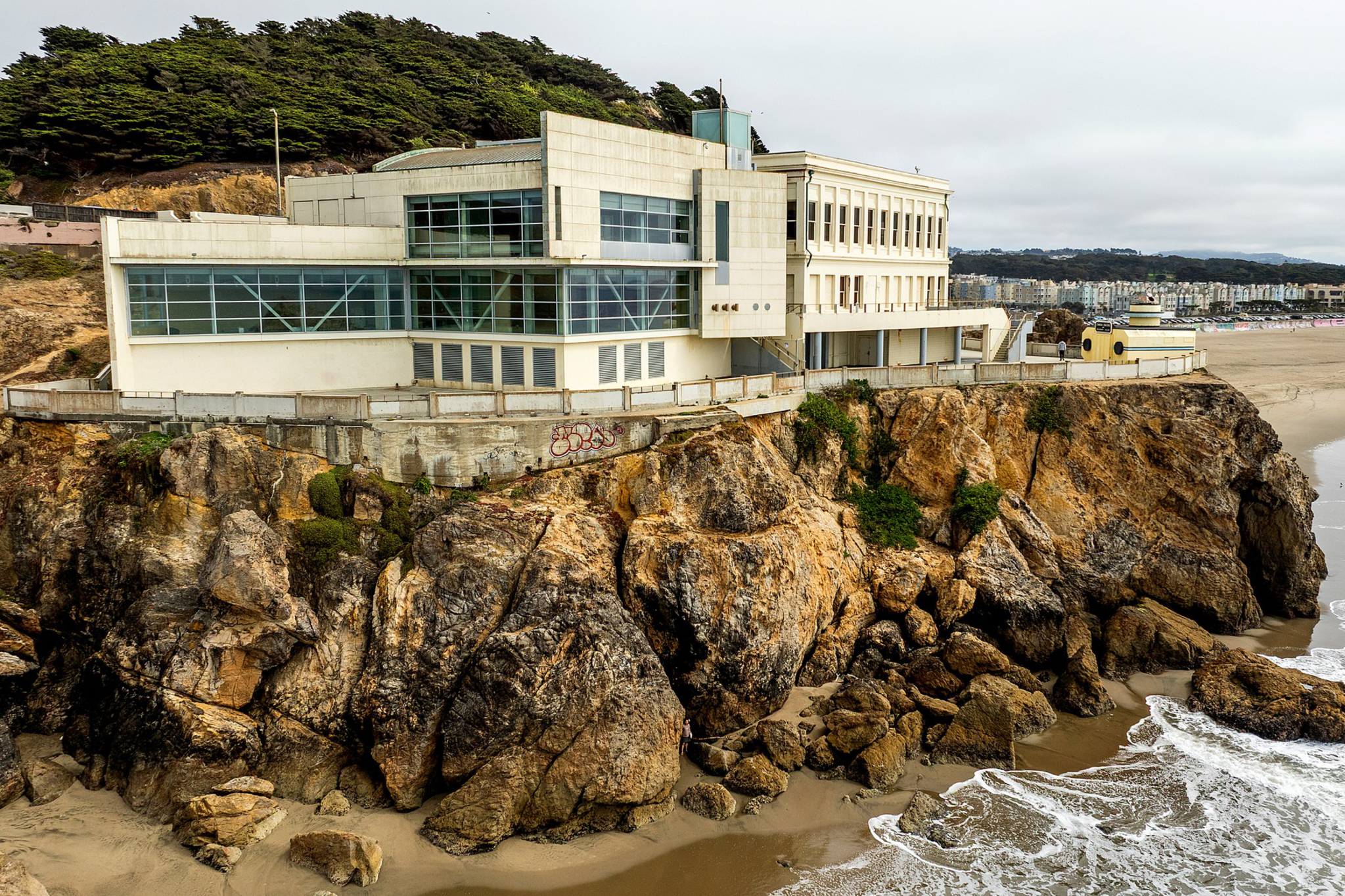 A large, white building sits atop rugged, rocky cliffs overlooking a sandy beach and ocean waves, with trees and cityscape in the background.