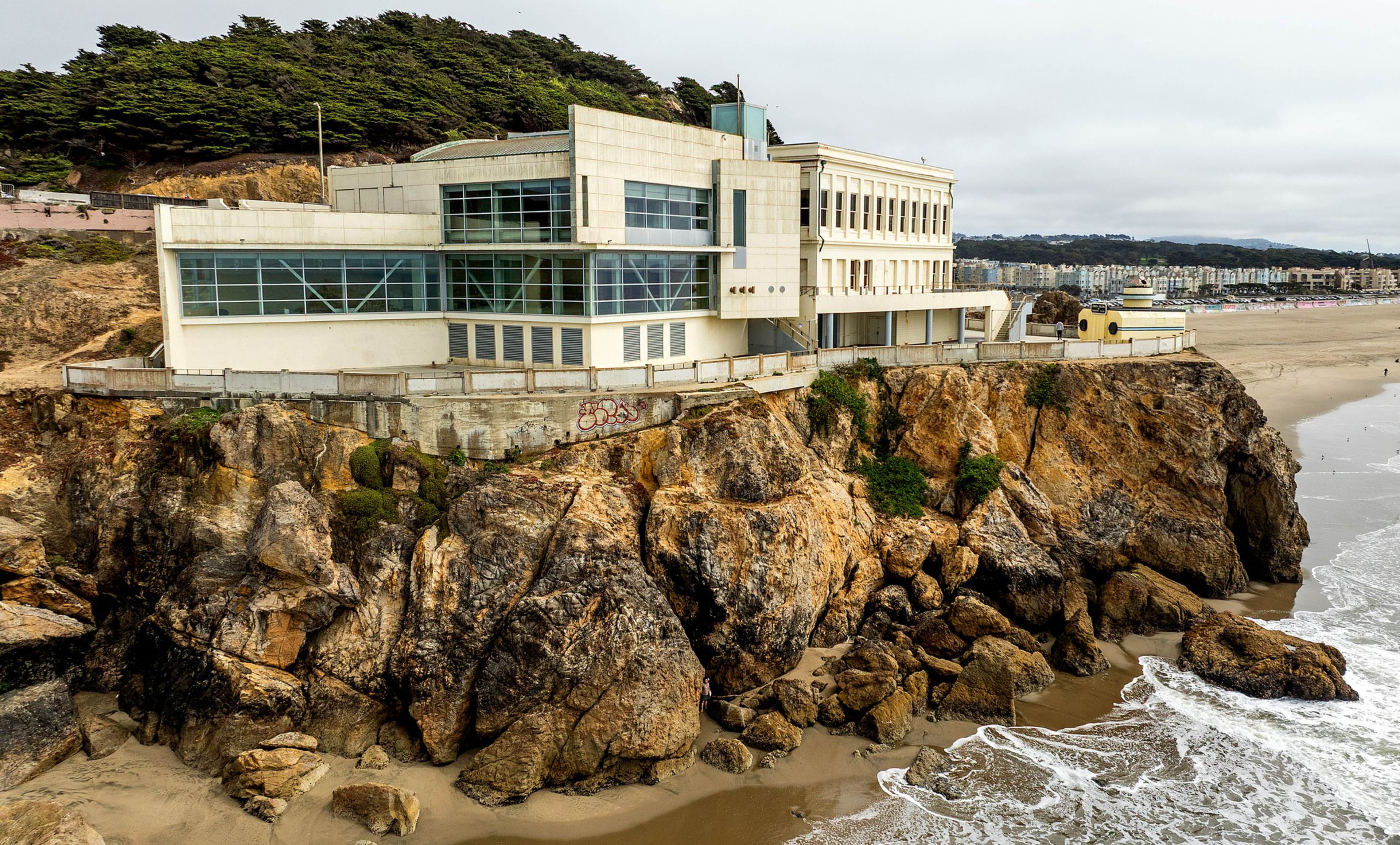 A large, white building sits atop rugged, rocky cliffs overlooking a sandy beach and ocean waves, with trees and cityscape in the background.