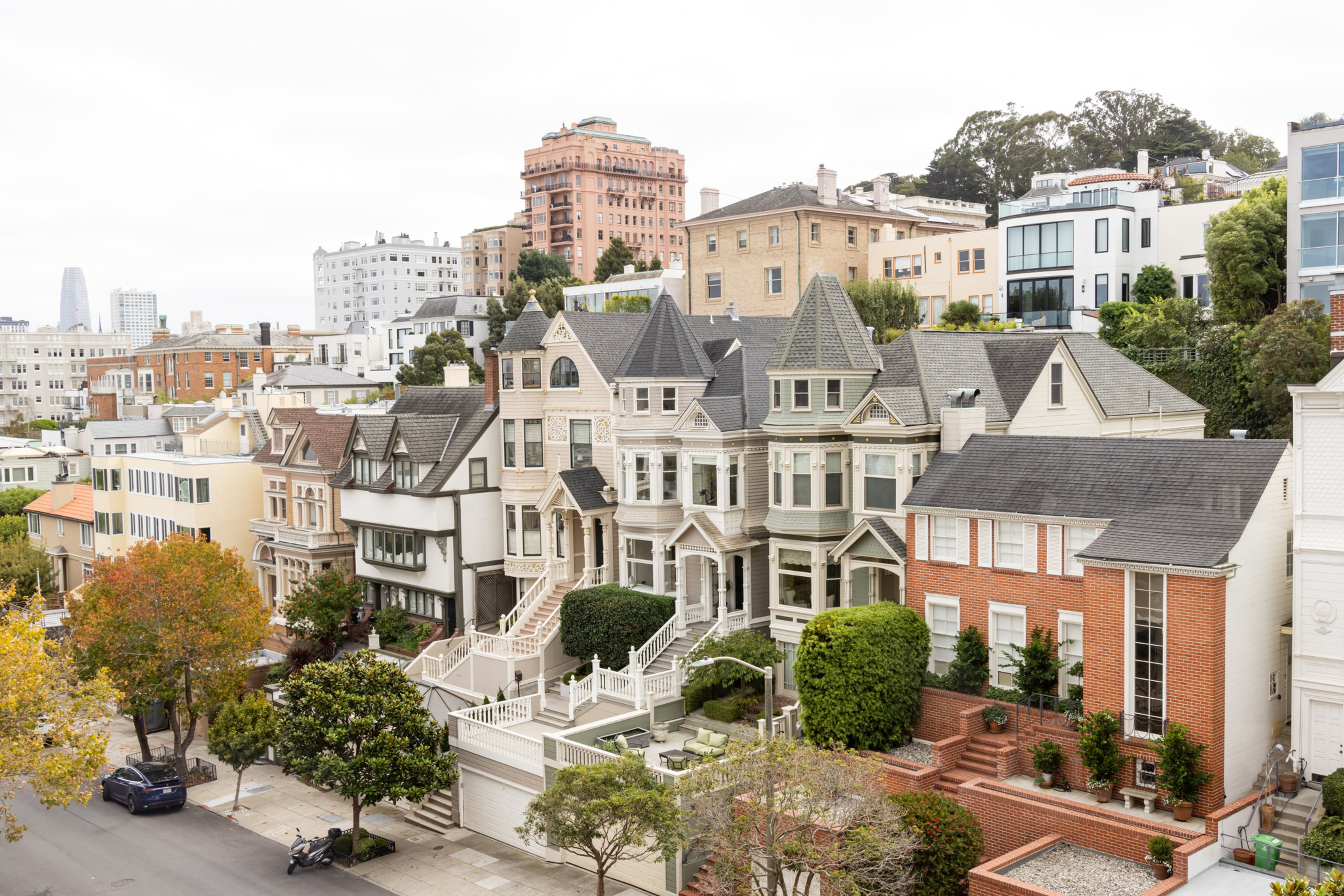 A row of colorful Victorian-style houses with steep roofs and bay windows lines a residential street, with urban buildings and trees in the background.
