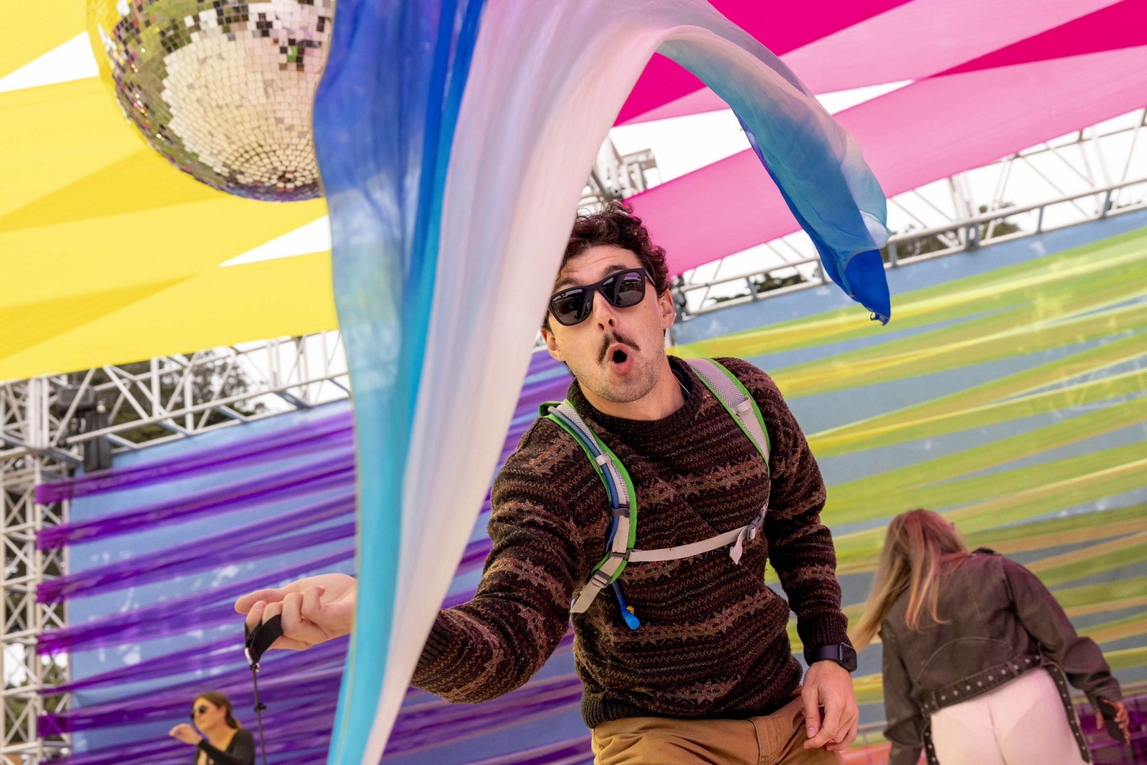 A man wearing sunglasses, a brown sweater, and a backpack waves blue and white fabric under a colorful canopy with a disco ball, with people in the background.
