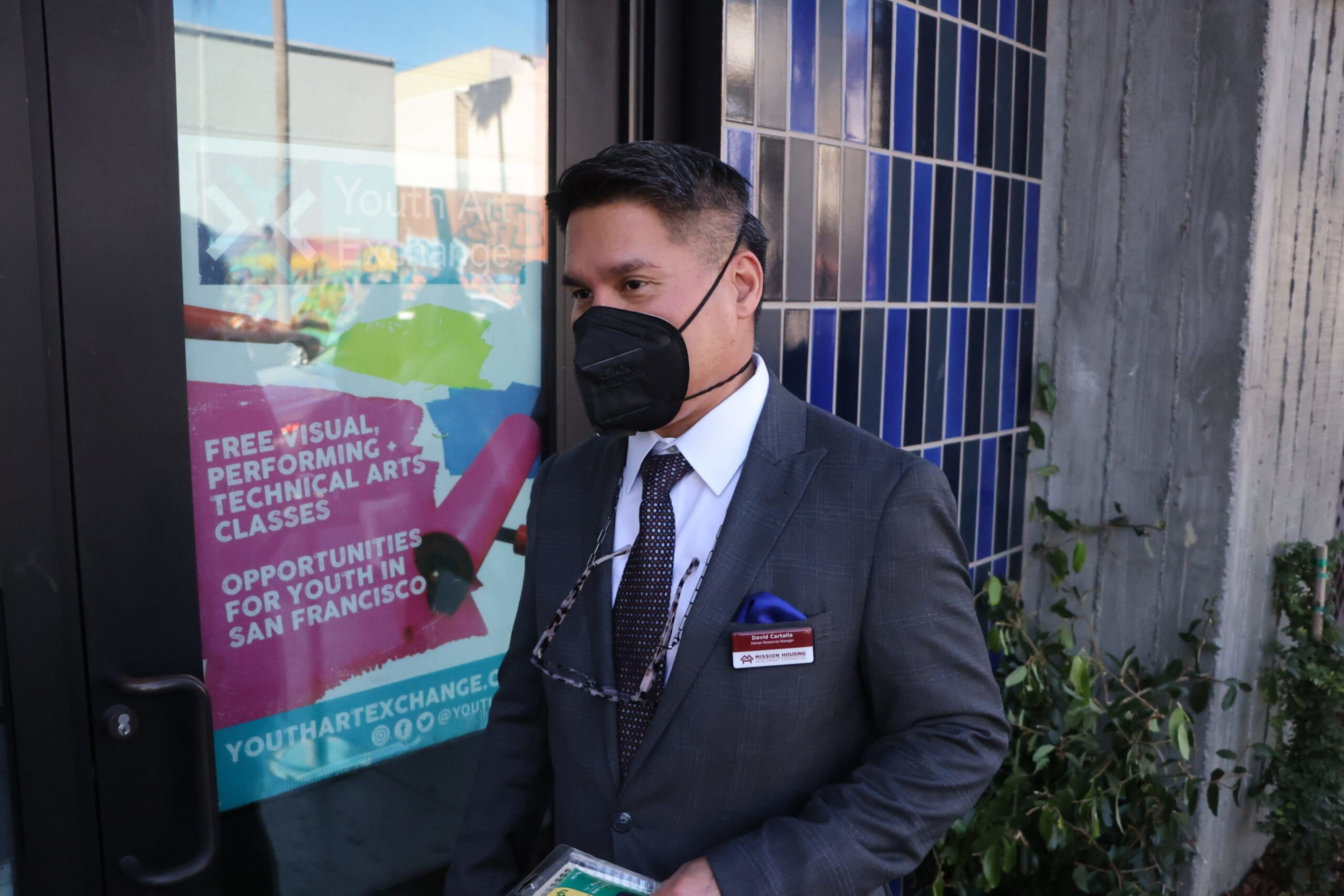 A man in a suit wearing a black mask and name tag stands near a door with a colorful poster about free arts classes for youth in San Francisco.