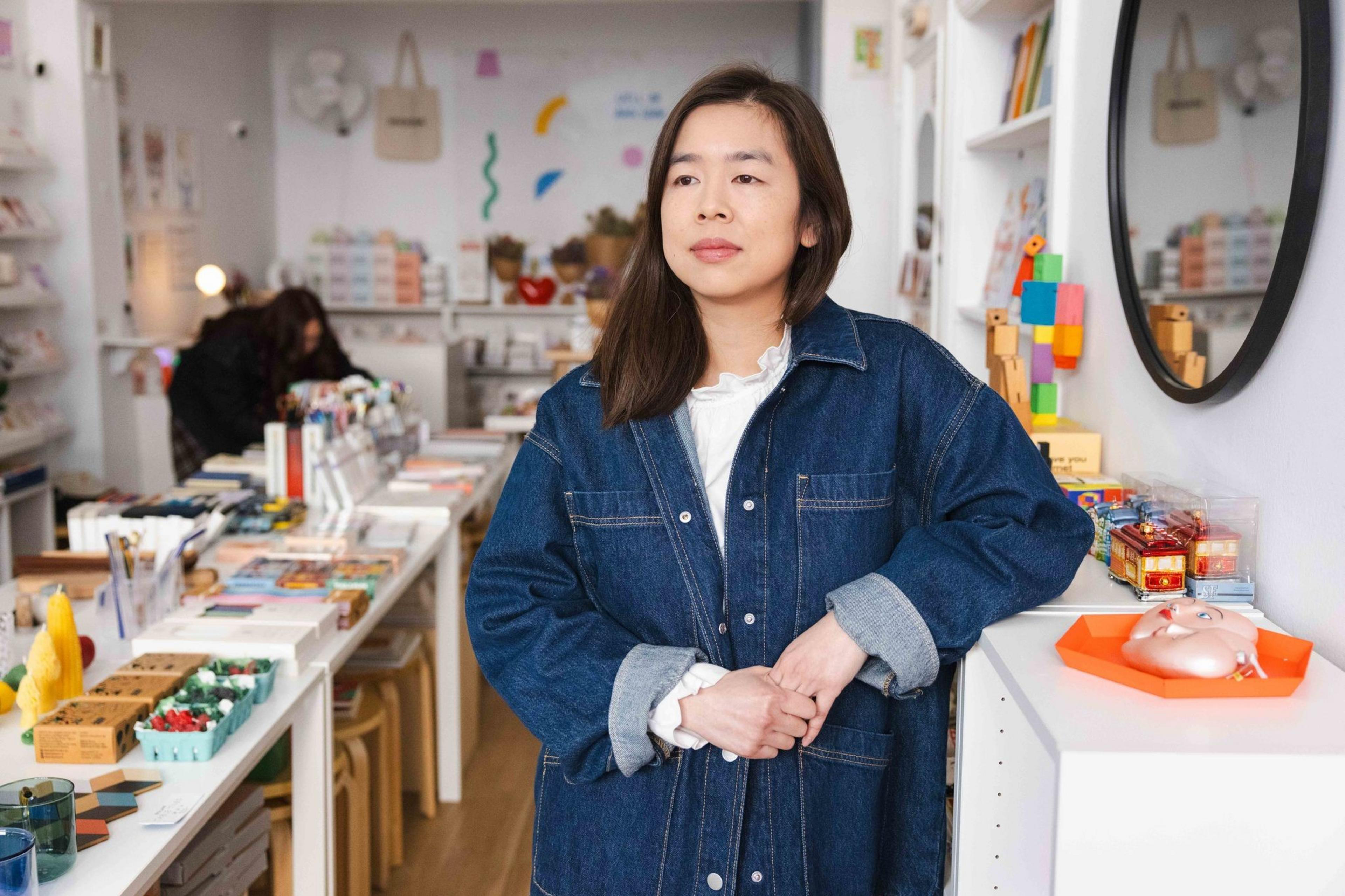 A person stands in a shop wearing a denim jacket, surrounded by colorful items on tables and shelves. The space is bright and arranged neatly.