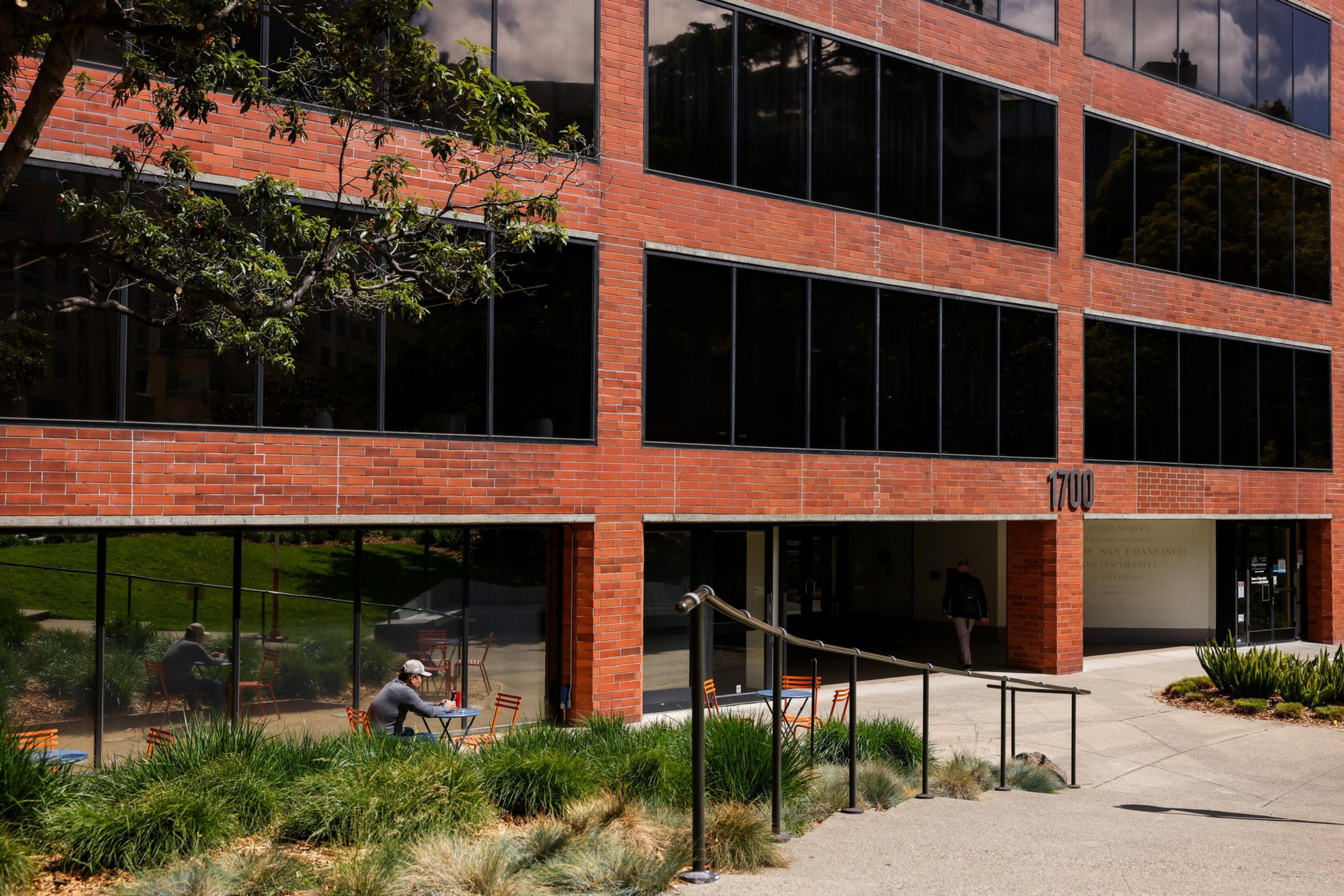 A modern brick building with dark windows stands behind a garden. A person sits at an outdoor table, and another walks into a shaded entrance labeled &quot;1700.&quot;
