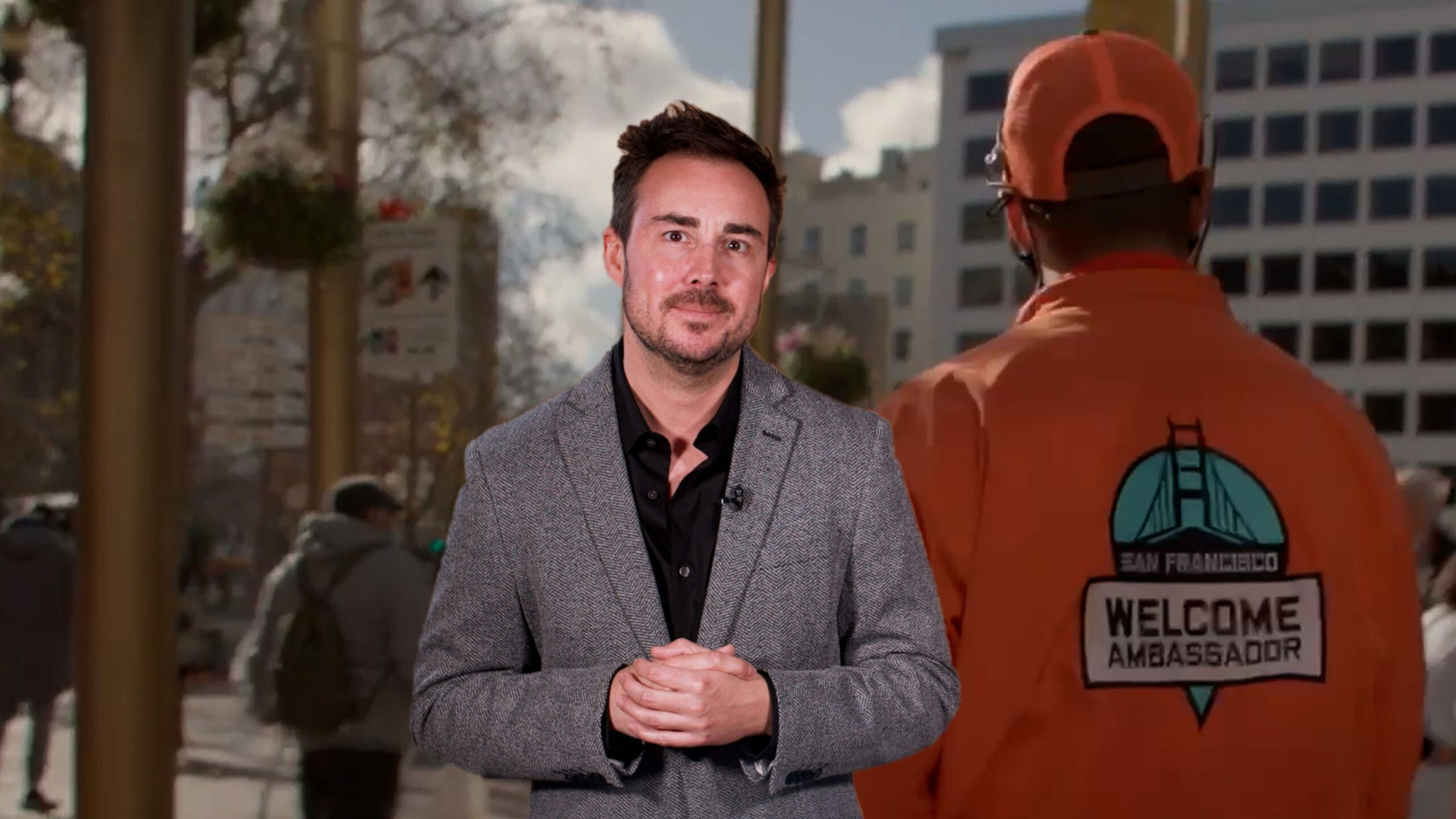 A man in a gray blazer stands facing the camera while another person in an orange jacket and cap with a "Welcome Ambassador" logo faces away.